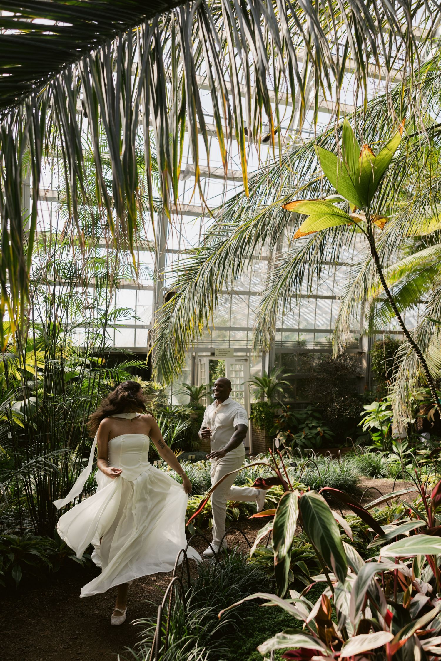 Two people in flowing white outfits dance through a greenhouse filled with palm fronds.