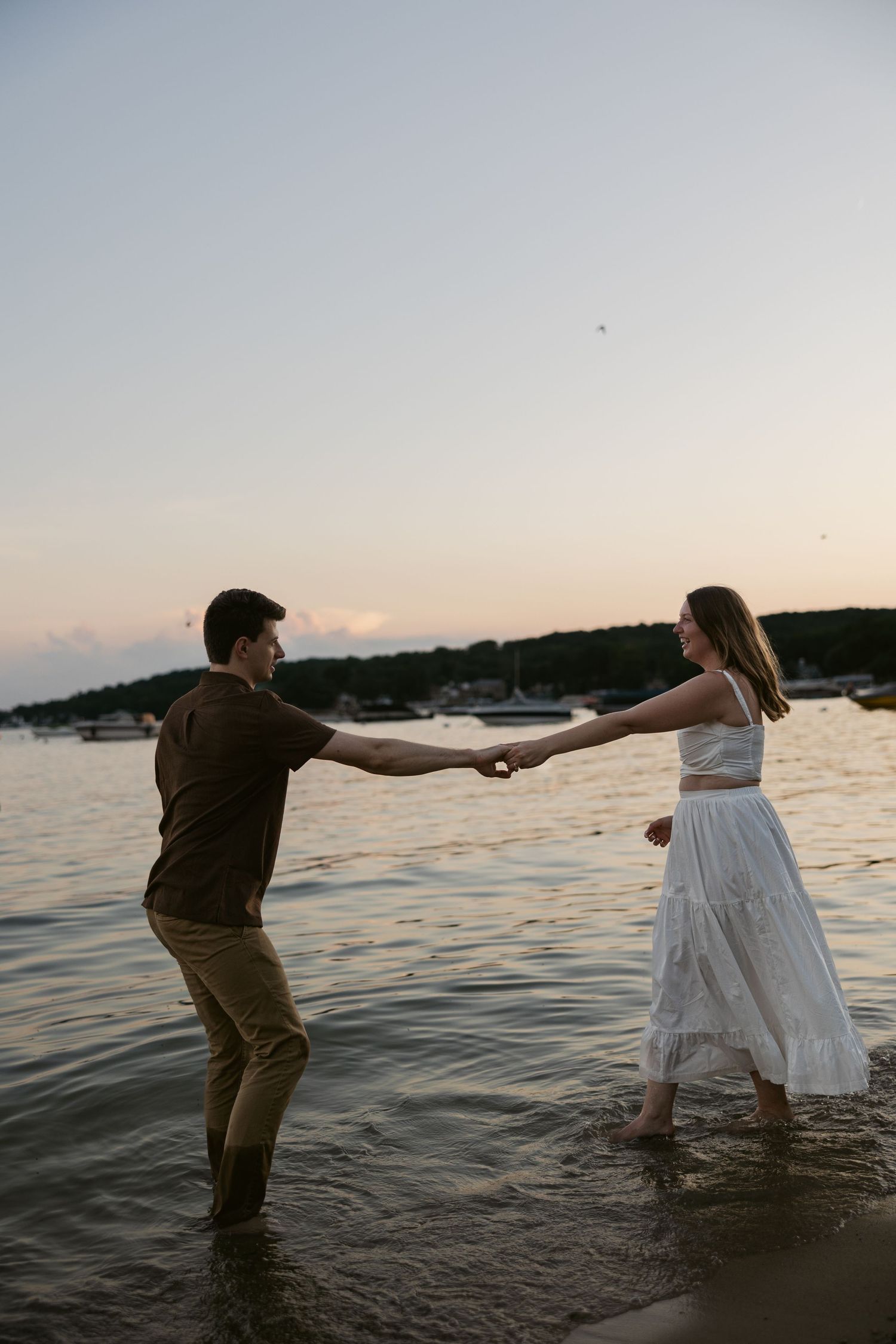 Two people playing and dancing in the ocean water during sunset with boats in the background.