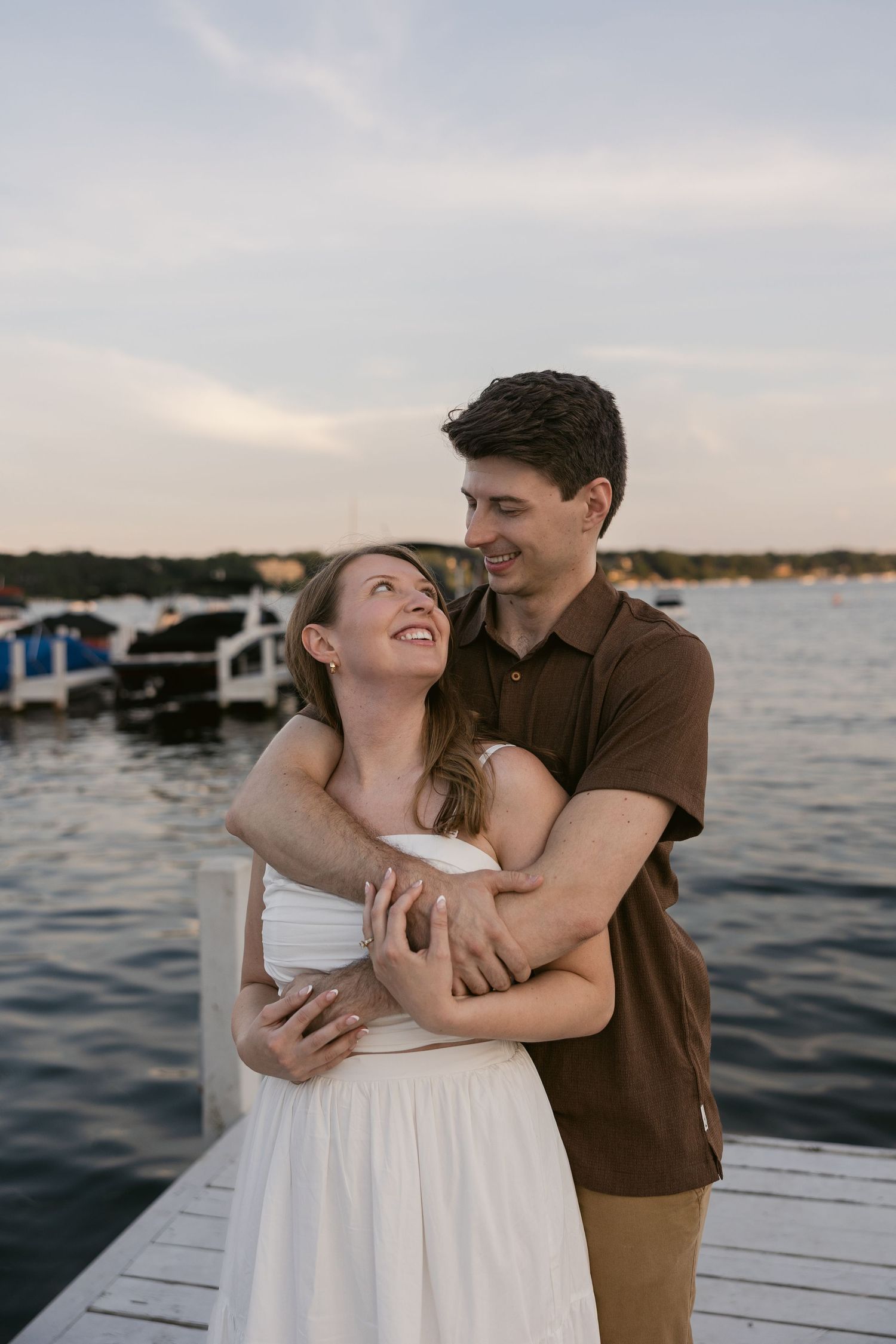 A couple shares a romantic moment on a dock by the water during sunset, embracing and laughing together.