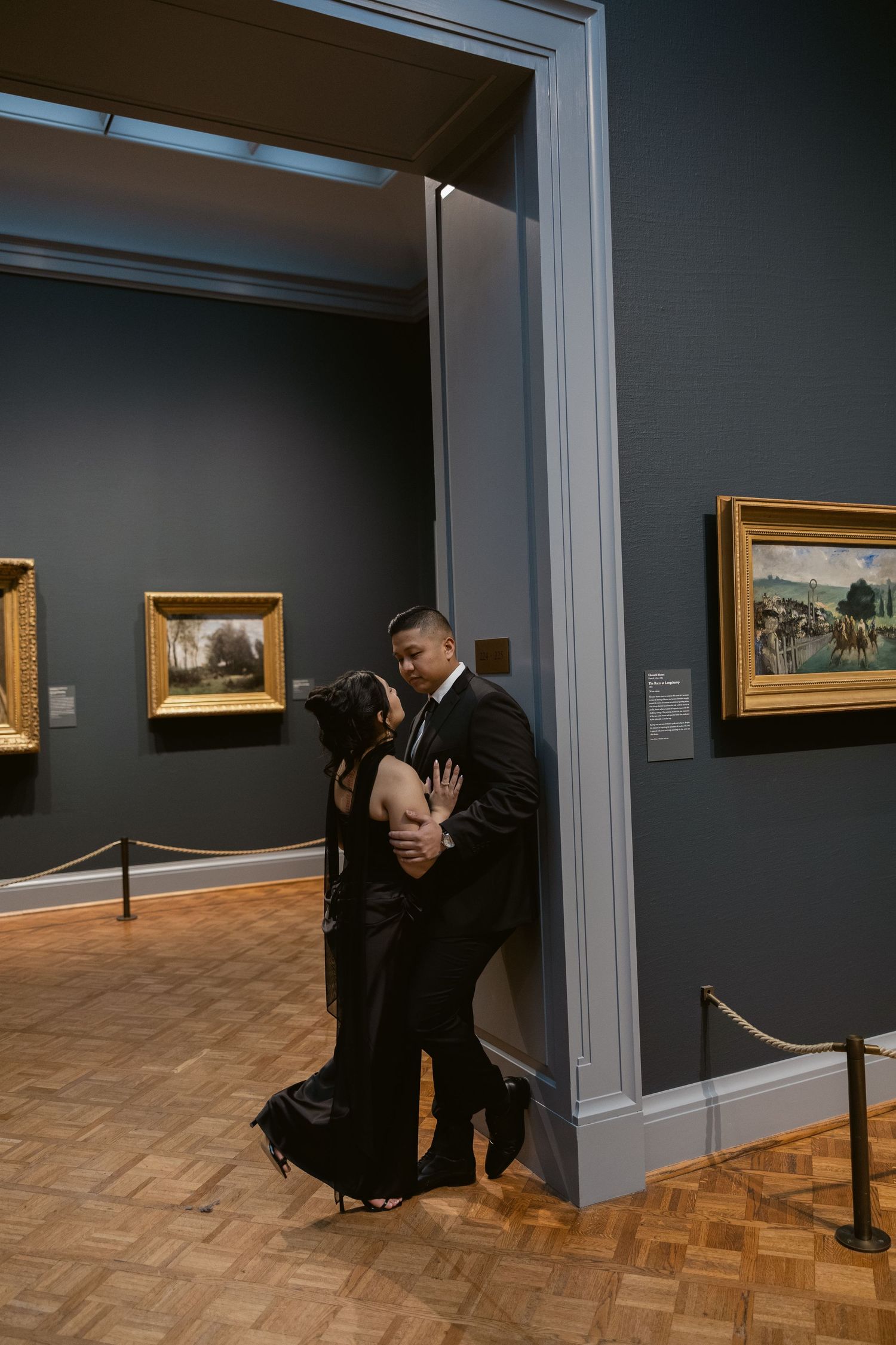 A couple poses intimately in an art gallery with framed paintings on dark gray walls.