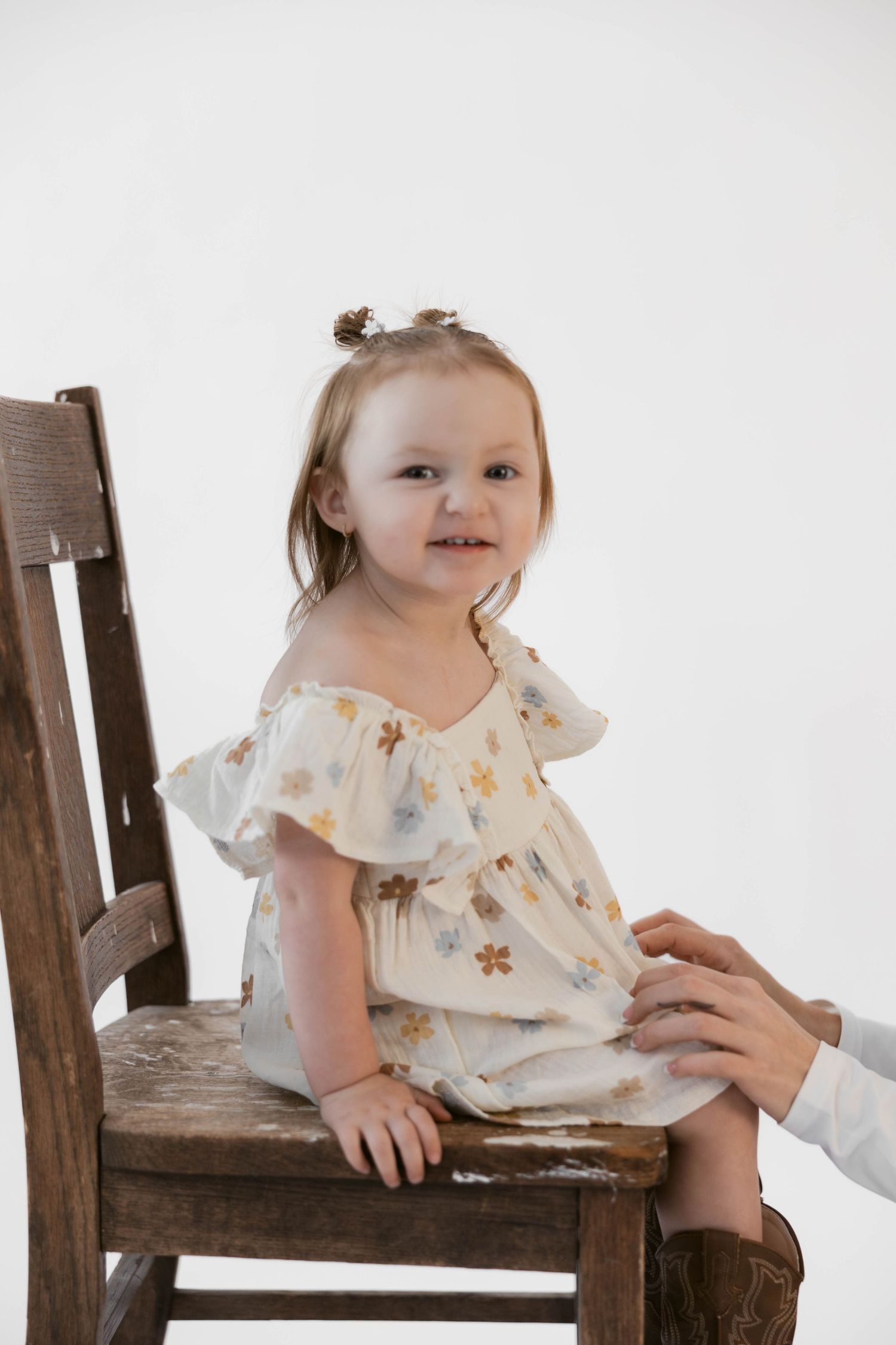 Toddler in floral dress and brown boots sits happily on wooden chair.