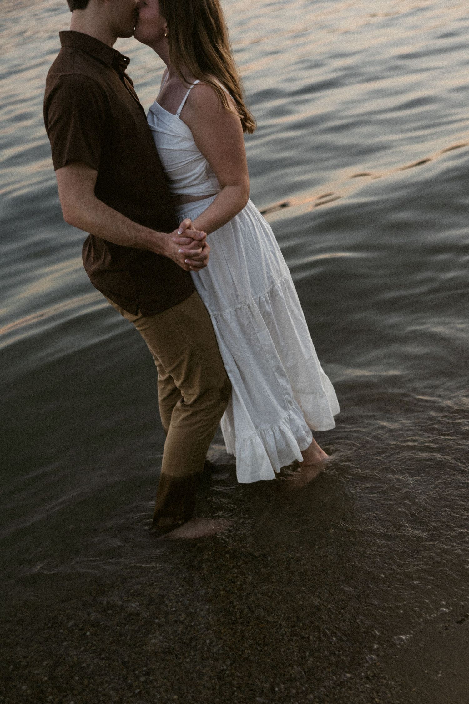 Couple in casual attire shares tender moments at the water's edge during sunset.