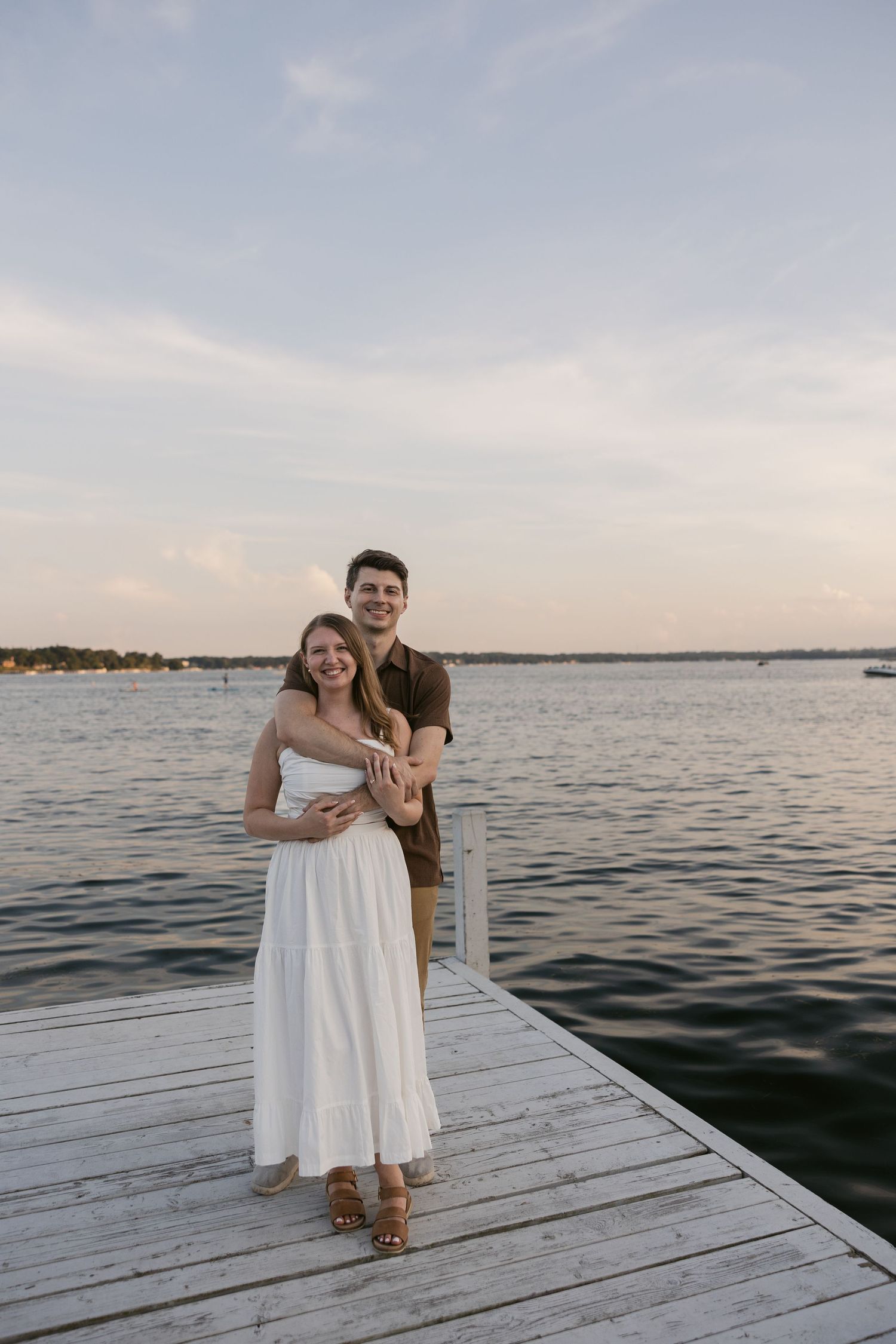 A couple shares a tender moment together on a white wooden dock overlooking calm waters at dusk.