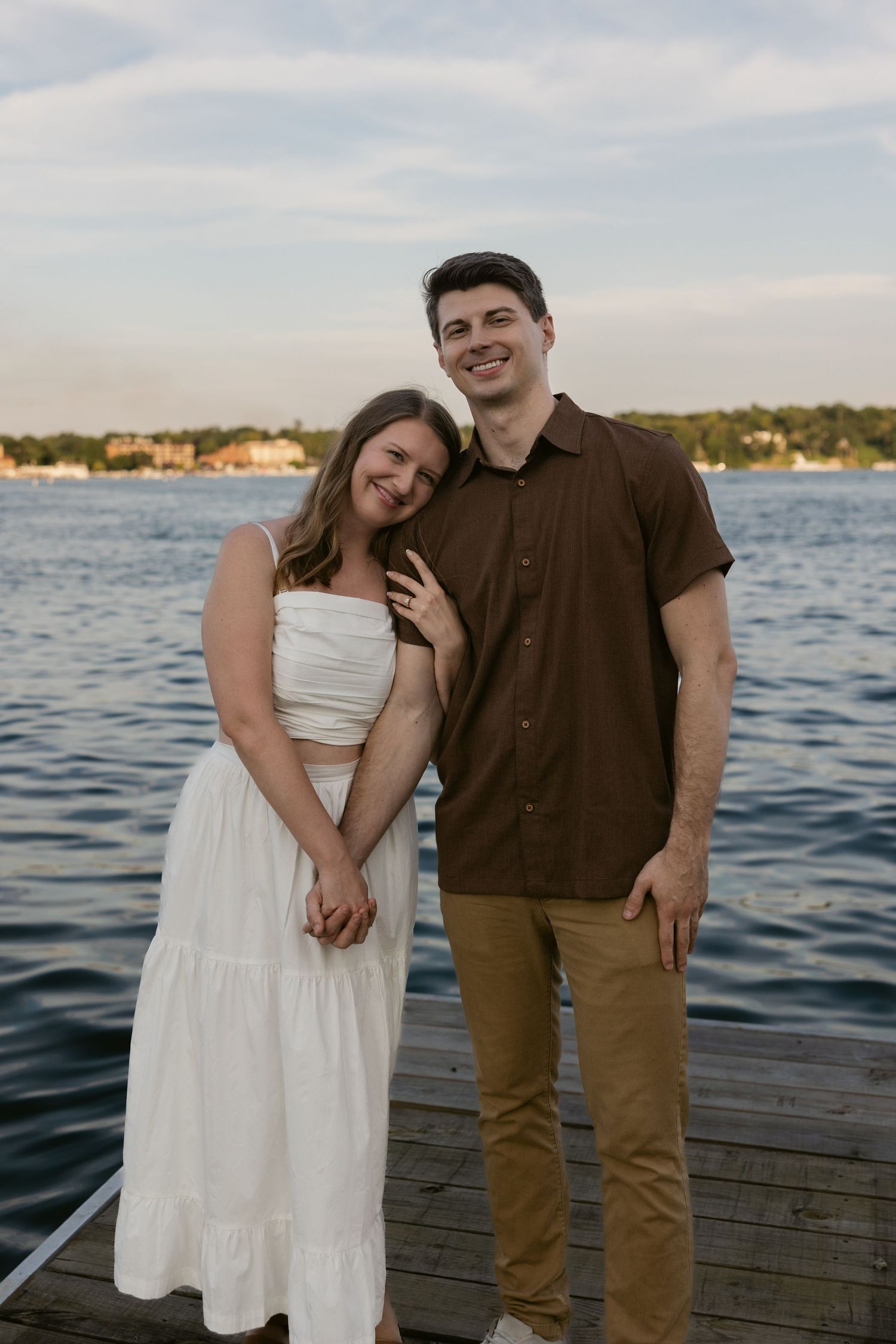 A couple stands together on a wooden dock overlooking a waterfront at sunset.