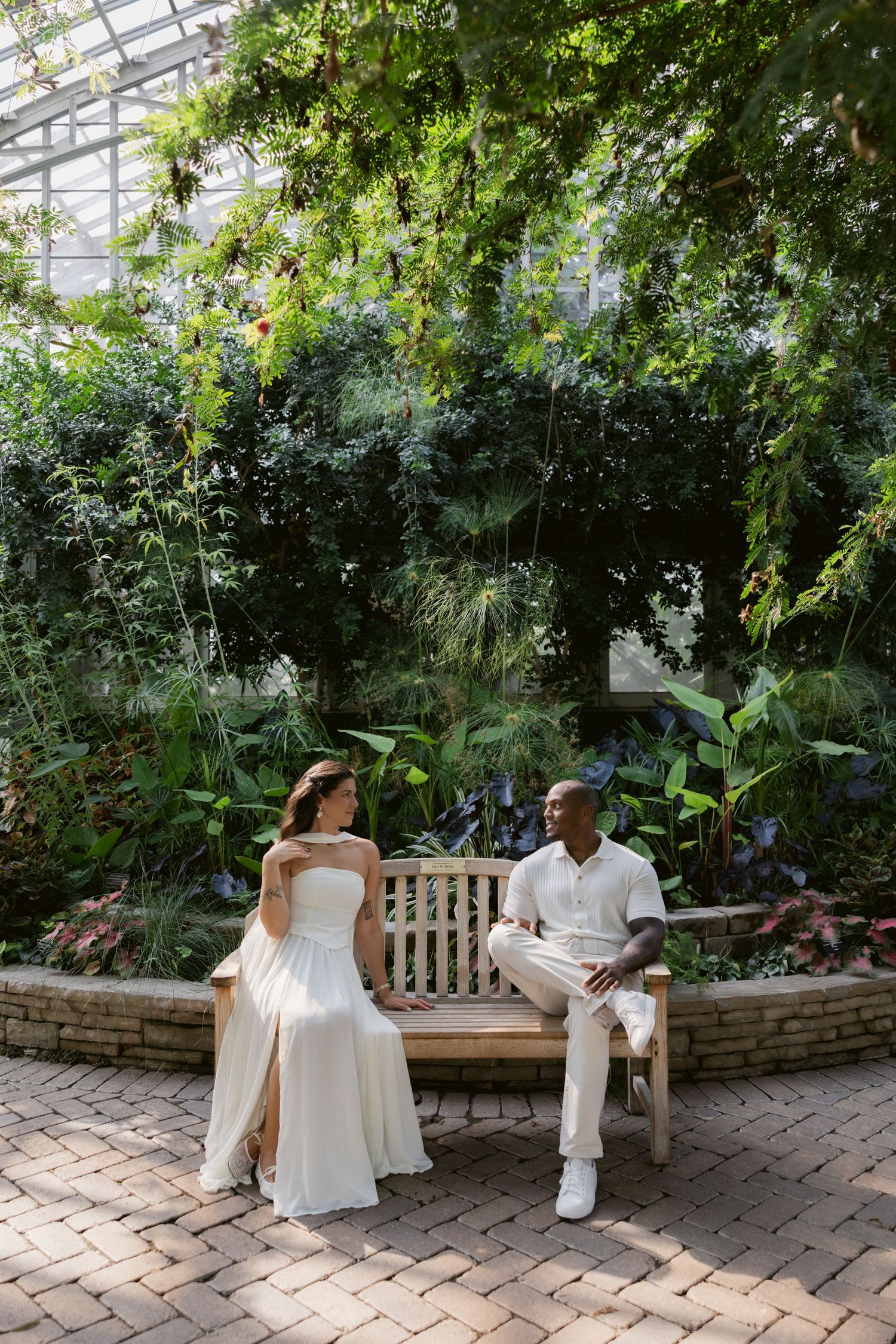 Couple sitting together on a wooden bench in a lush indoor greenhouse setting.