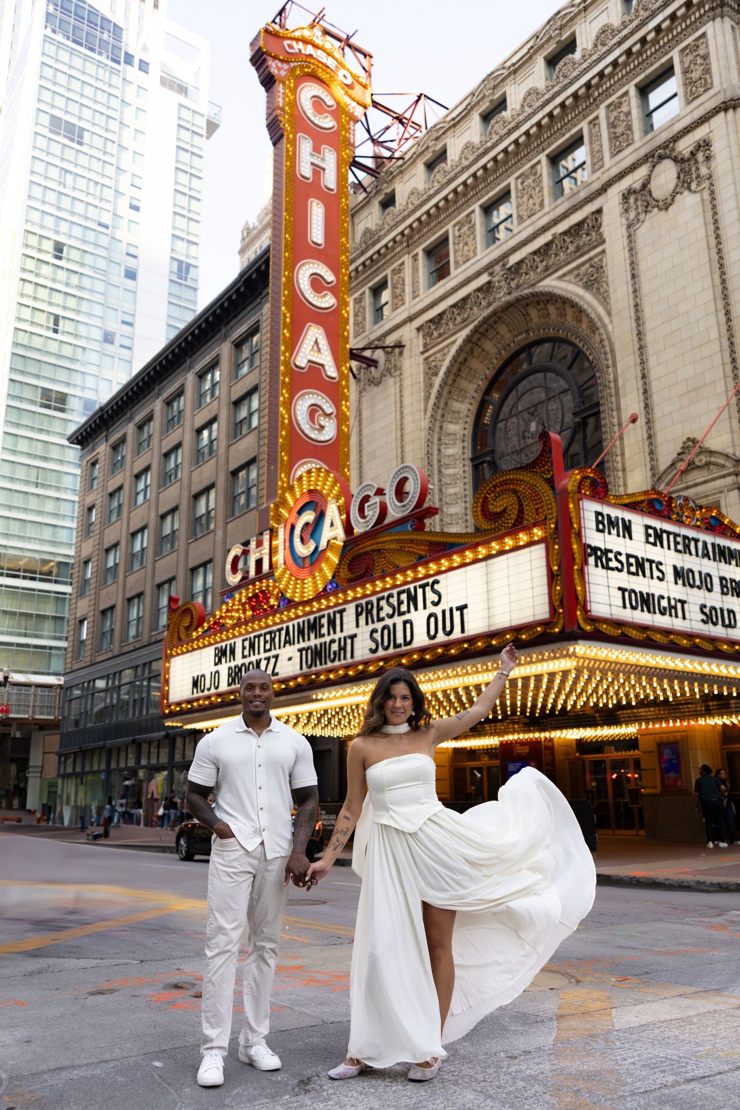 Dancing couple in white outfits pose playfully in front of the illuminated Chicago Theatre sign.