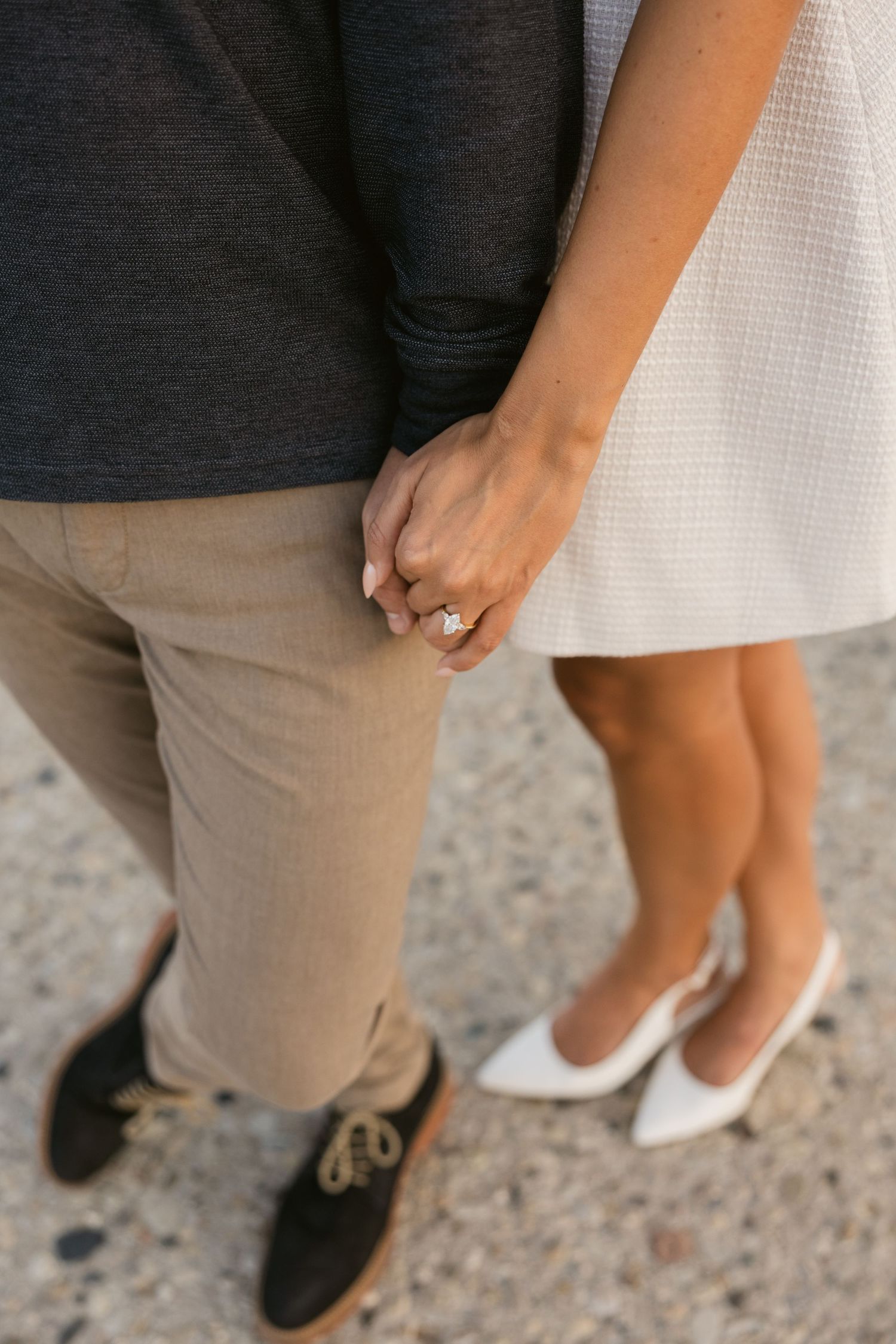 Close-up of holding hands with engagement ring against khaki pants and white dress with contrasting footwear.