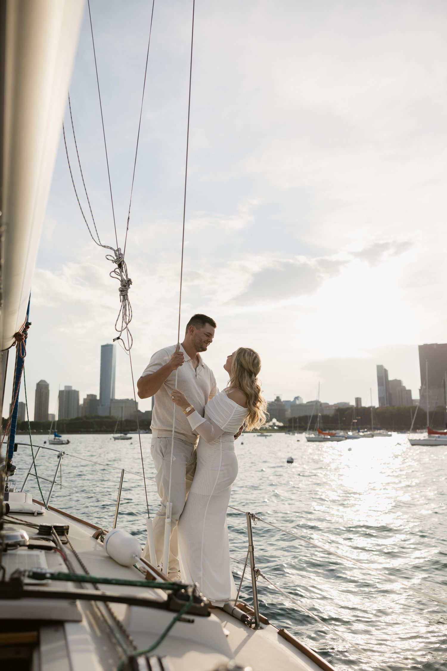 Couple in white outfits sharing an intimate moment on a sailboat with city skyline at sunset.