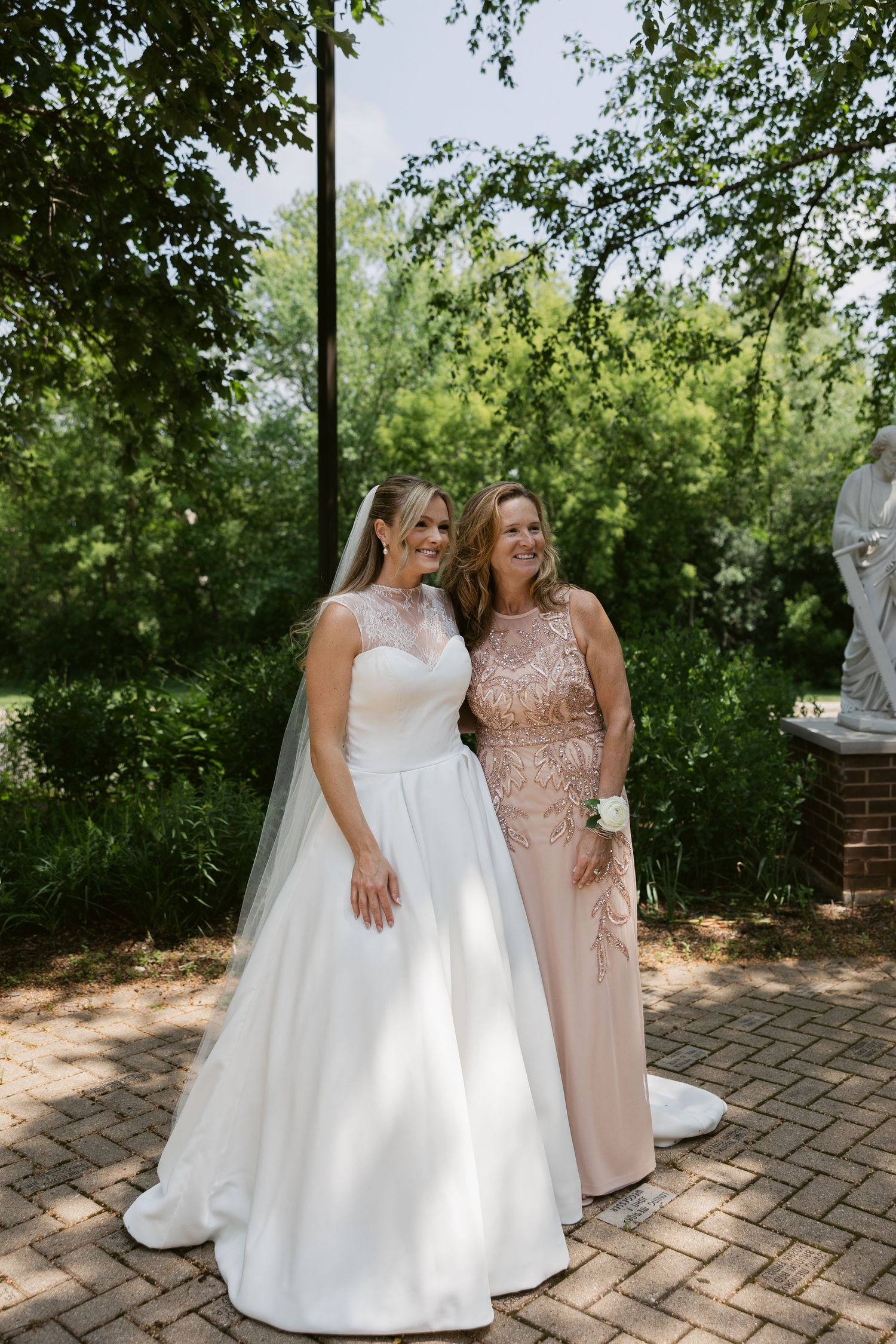Two women pose for a formal photo on brick pathway surrounded by greenery.