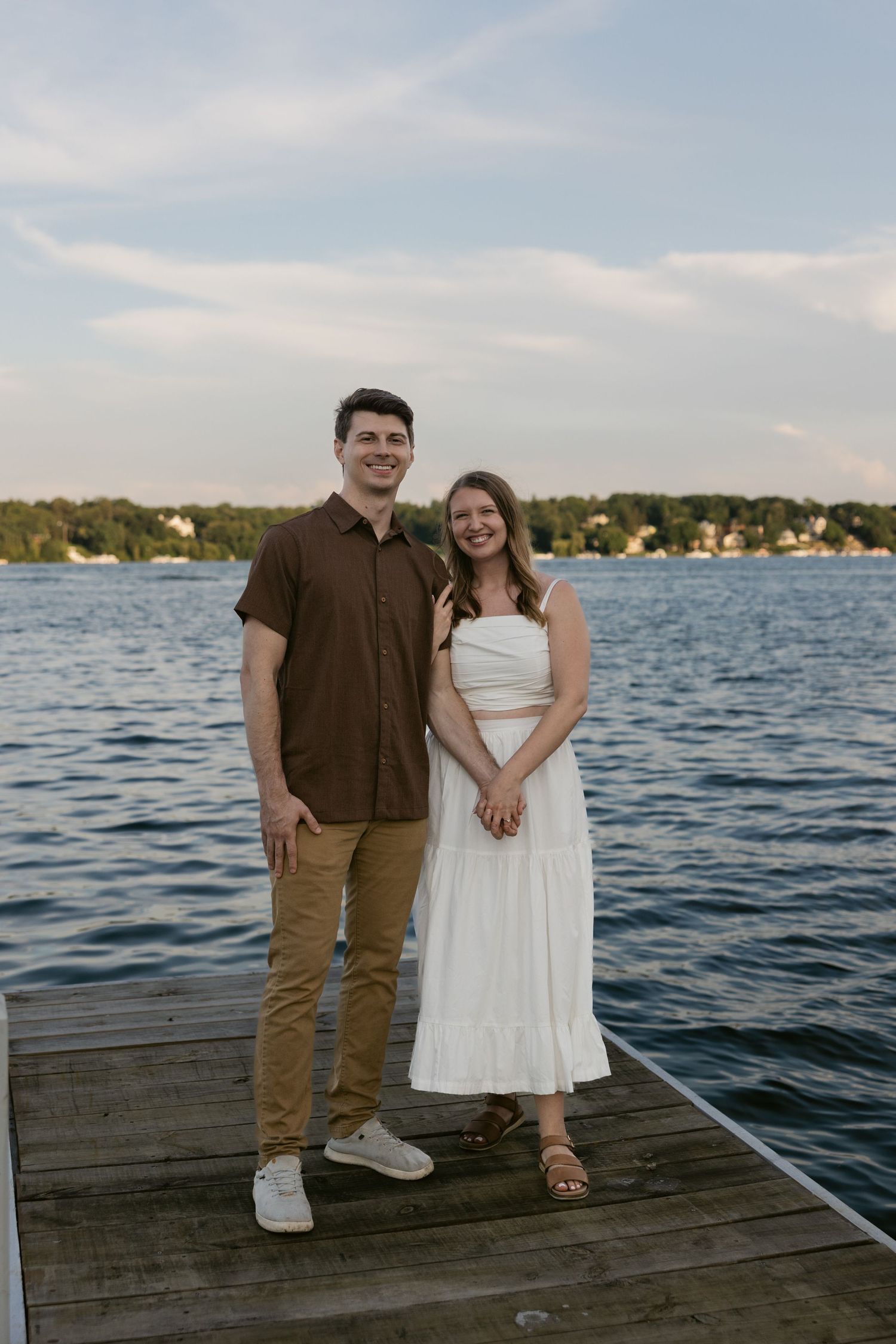 A couple stands on a wooden dock at sunset overlooking a peaceful lake with houses visible along the shoreline.
