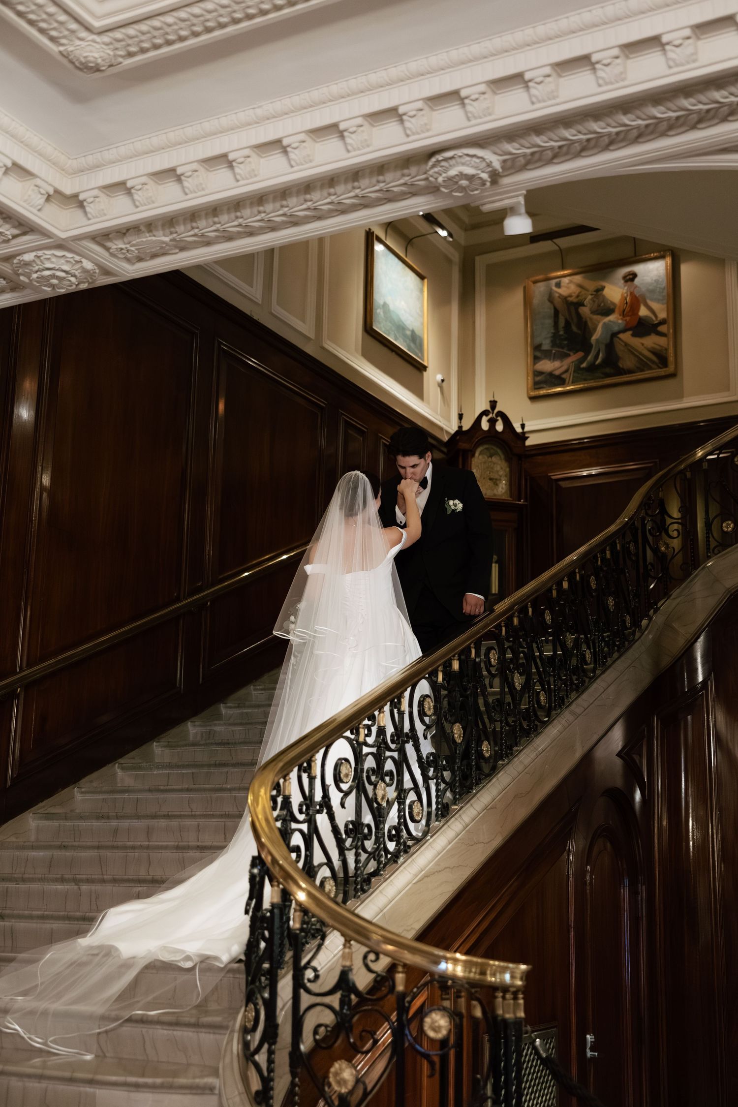 Elegant wedding dress flowing down a grand staircase with ornate iron railings and wood paneling in a historic venue.