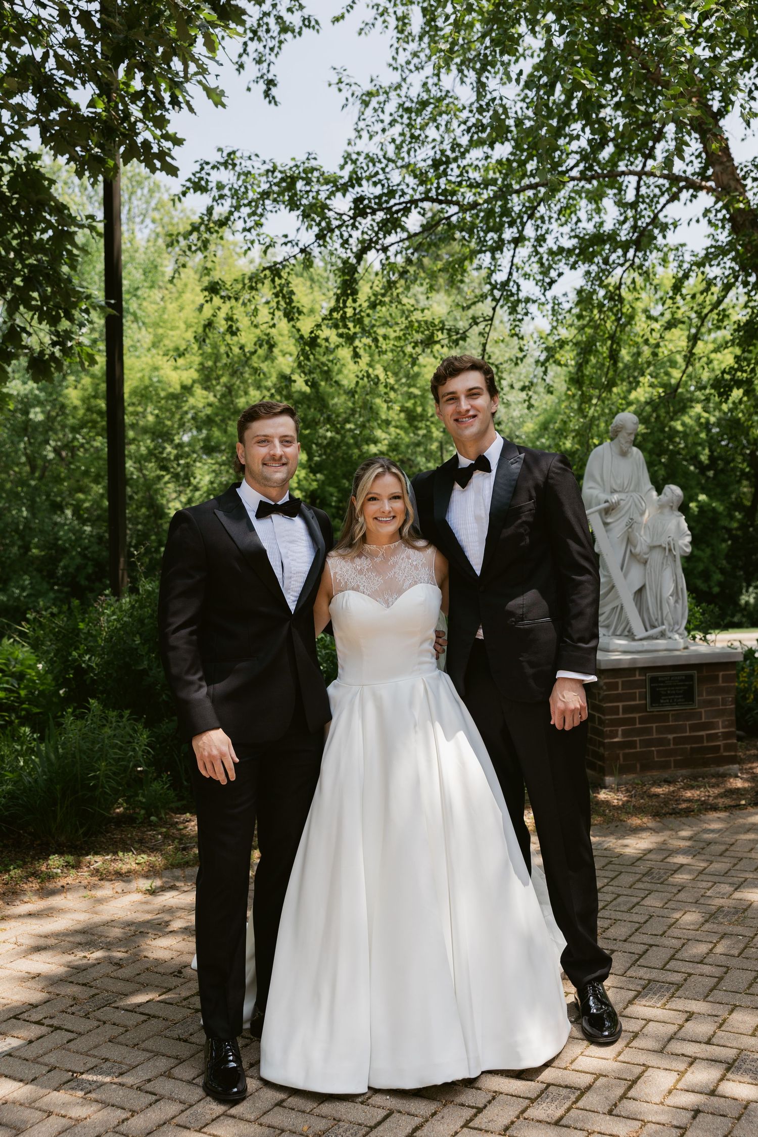 A wedding party poses on a brick path in a garden setting with elegant formal attire.