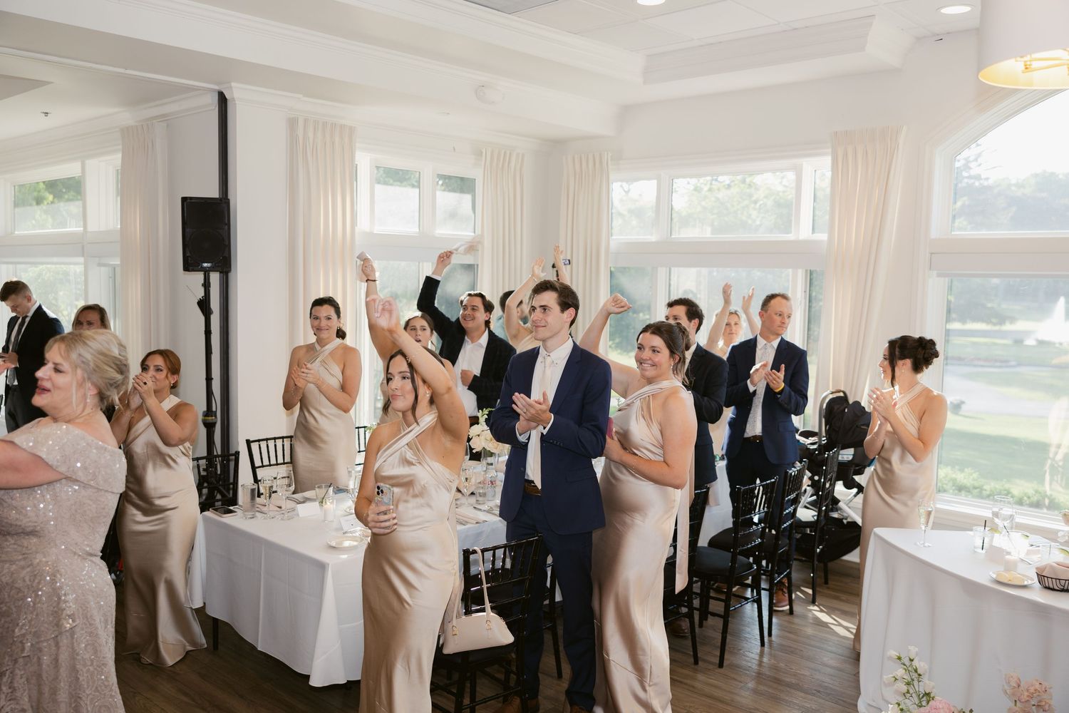 Wedding reception party gathers in an elegant white indoor venue with large windows.