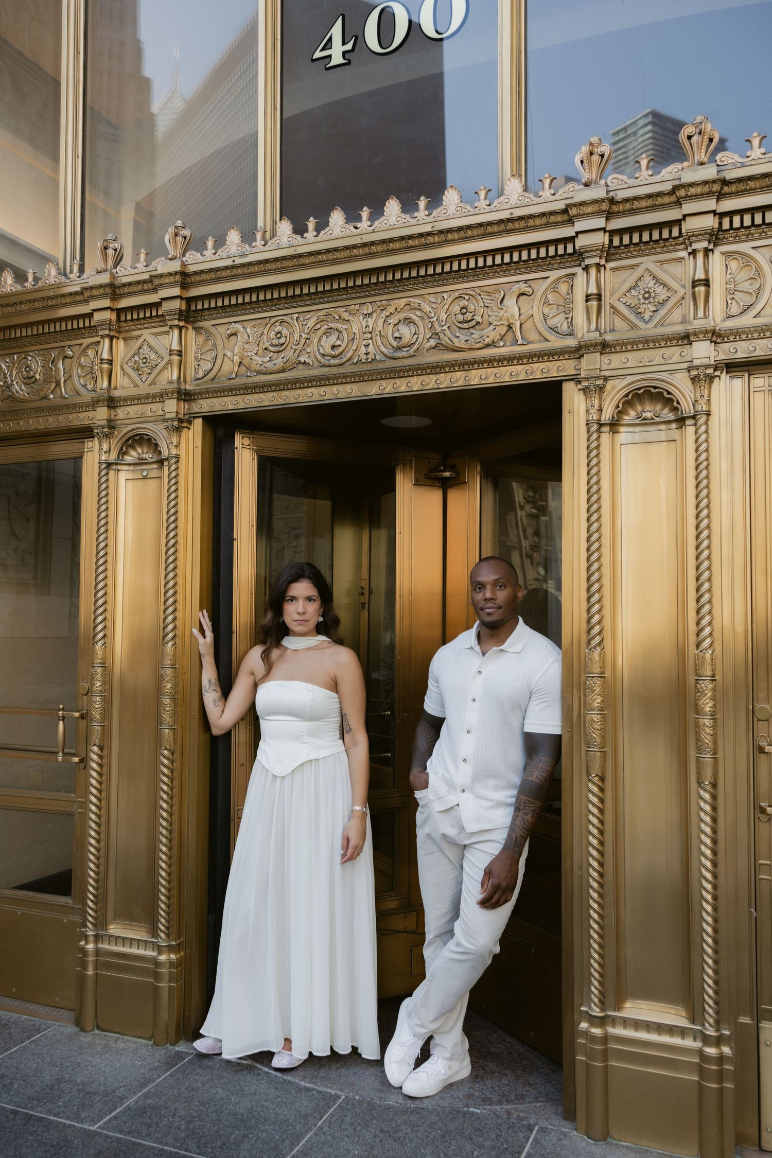 Couple in matching white outfits pose together in front of ornate gold elevator doors with number 400 above.
