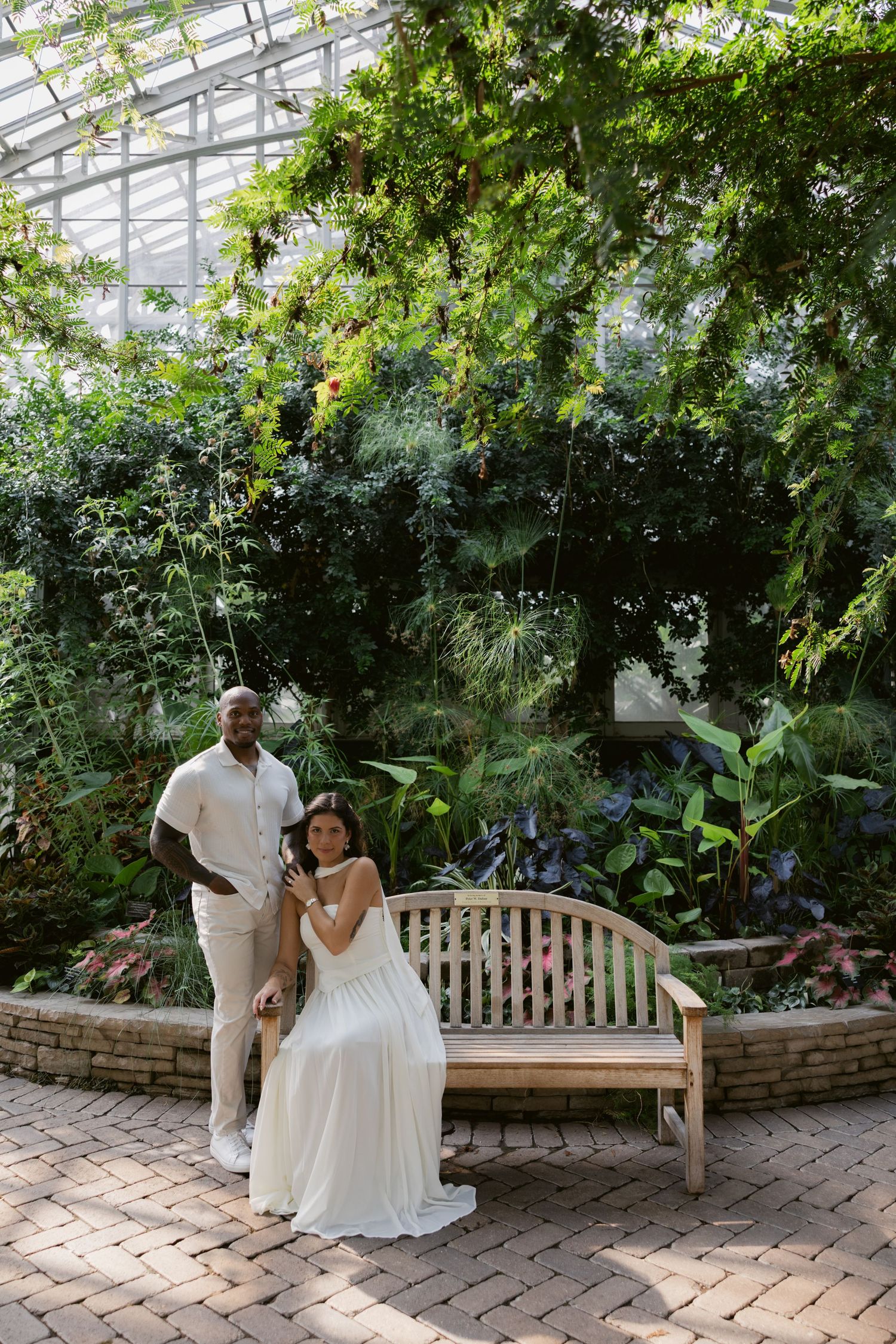 A couple shares an intimate moment on a wooden bench surrounded by greenery and brick walls.