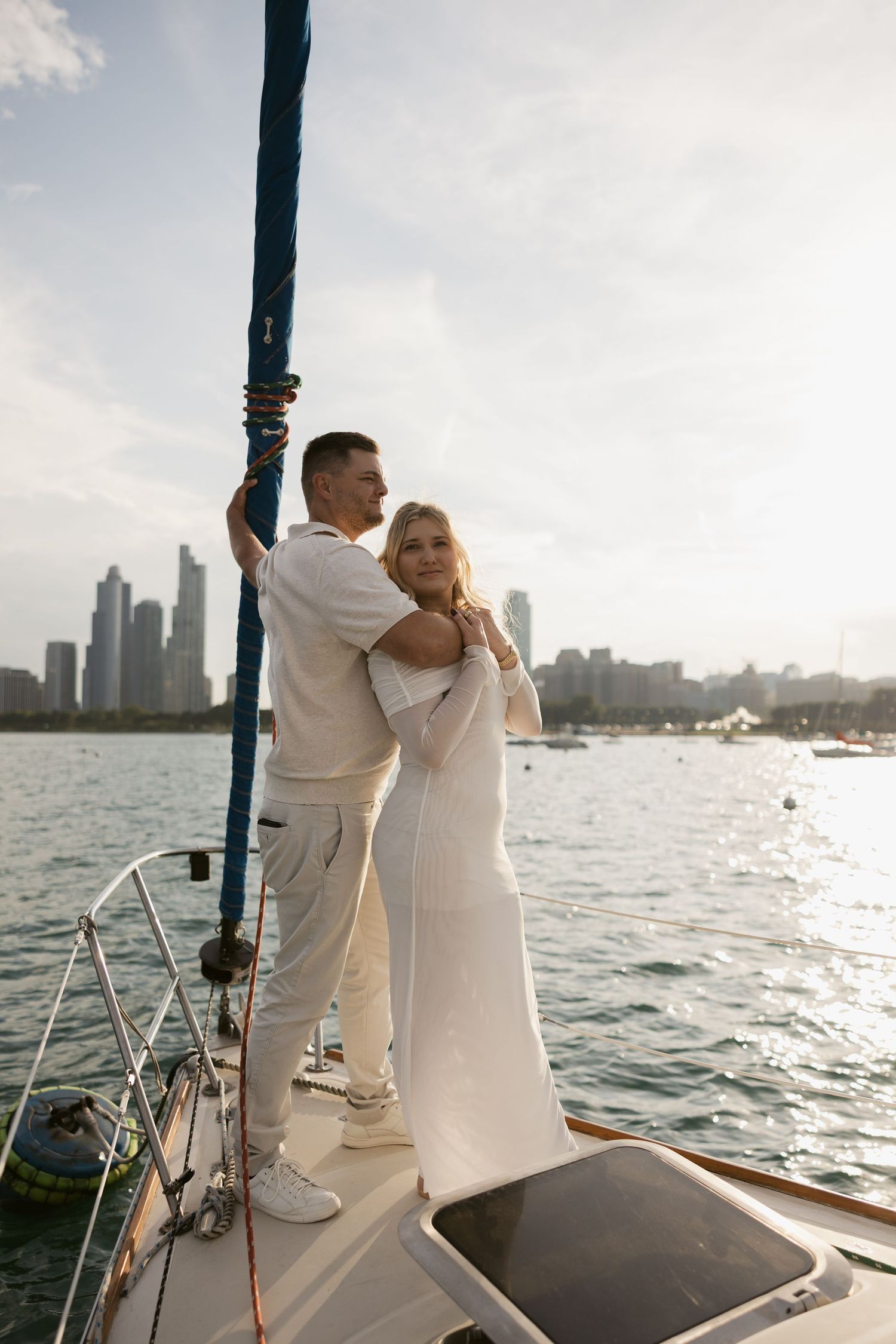 Couple dressed in white embracing on sailboat deck against waterfront cityscape during golden hour.