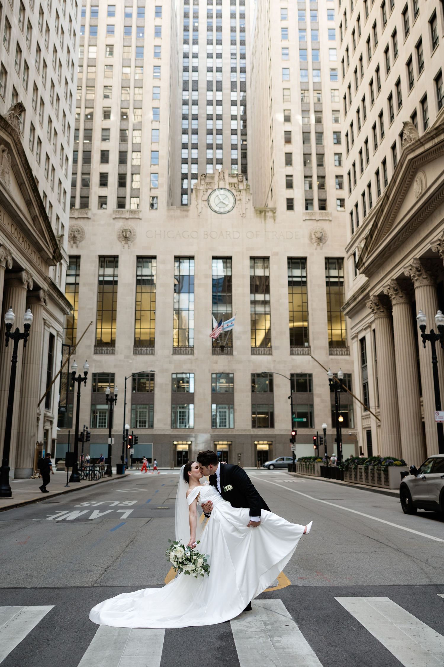 A bride's flowing veil creates a dramatic spiral motion as she twirls on a grand city street lined with tall buildings.