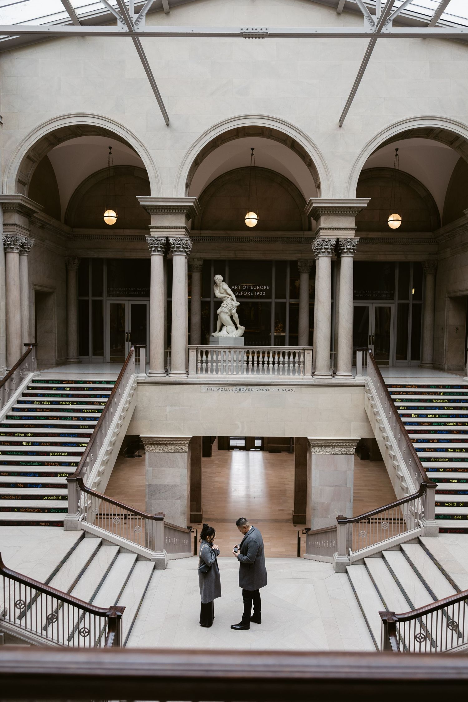 Two people stand in a grand marble building with arched columns and staircases.