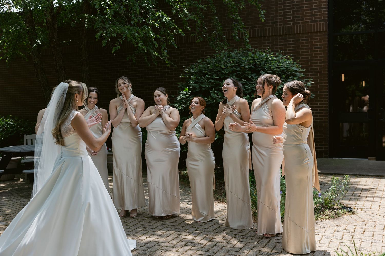 Bridal party in neutral champagne dresses reacting with joy and surprise on a brick path during wedding photos.