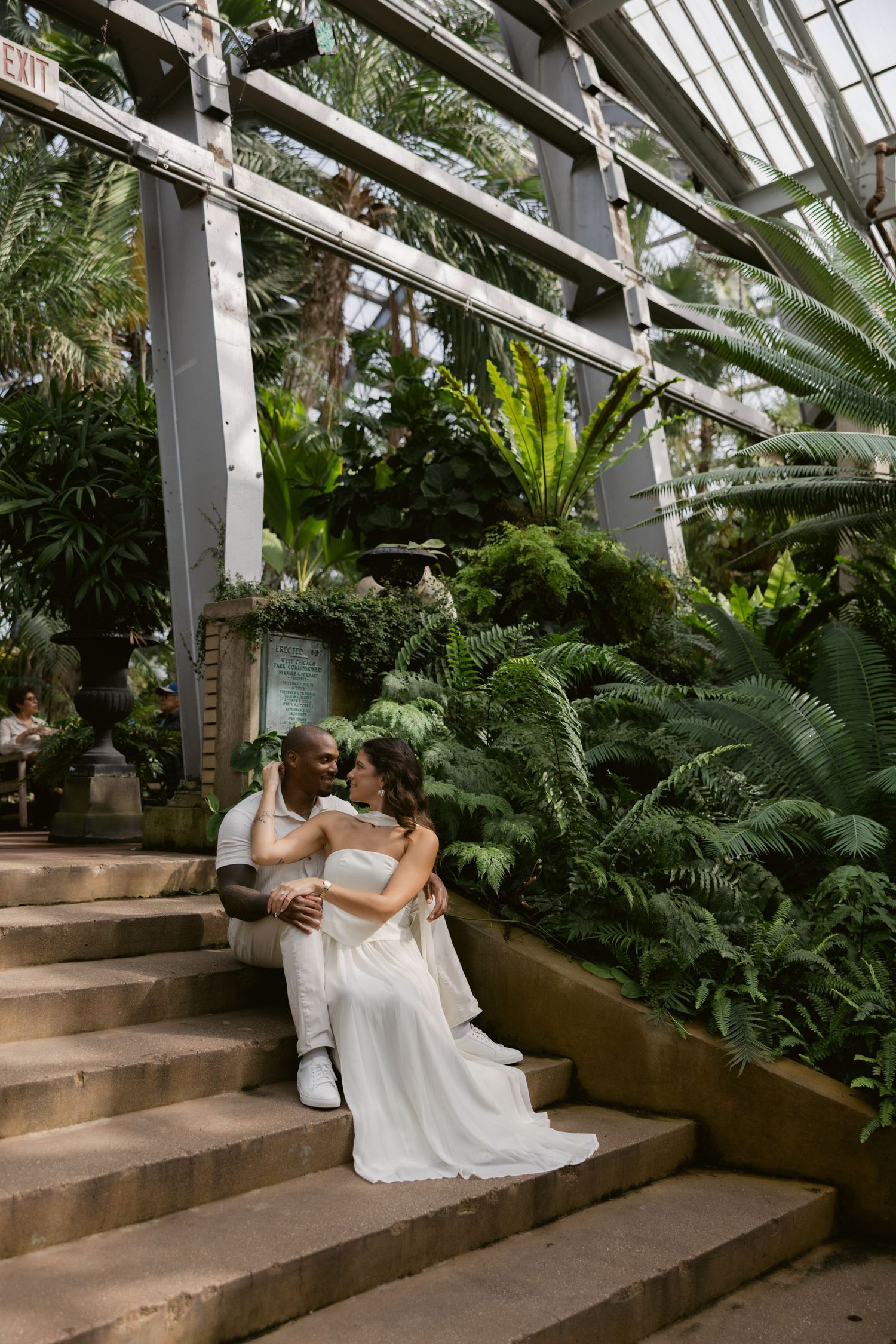 A couple in elegant white outfits sits together on stone steps inside a sunlit greenhouse surrounded by tropical plants.