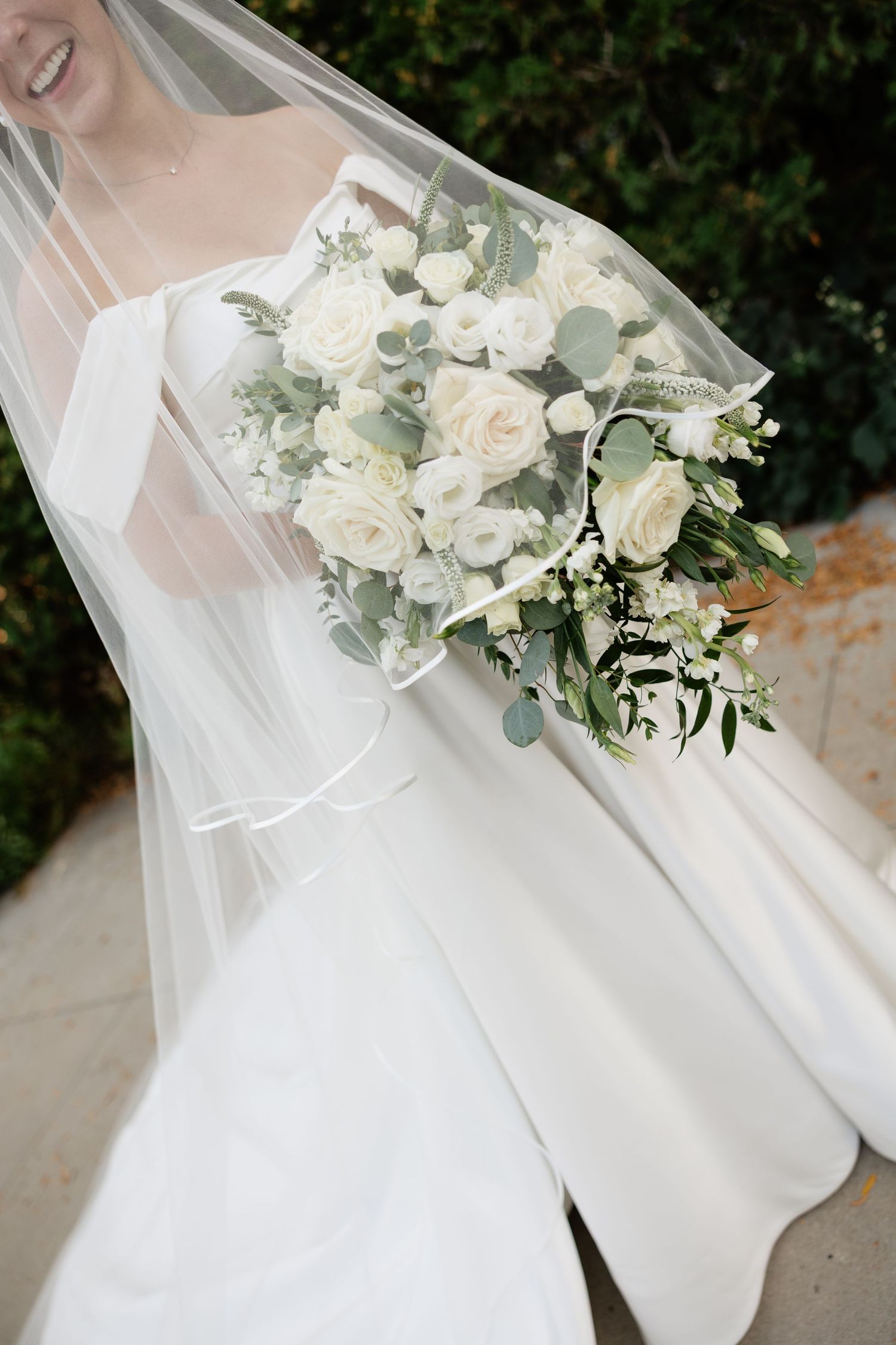 A delicate white bridal bouquet with roses and eucalyptus leaves against flowing white wedding dress fabric.