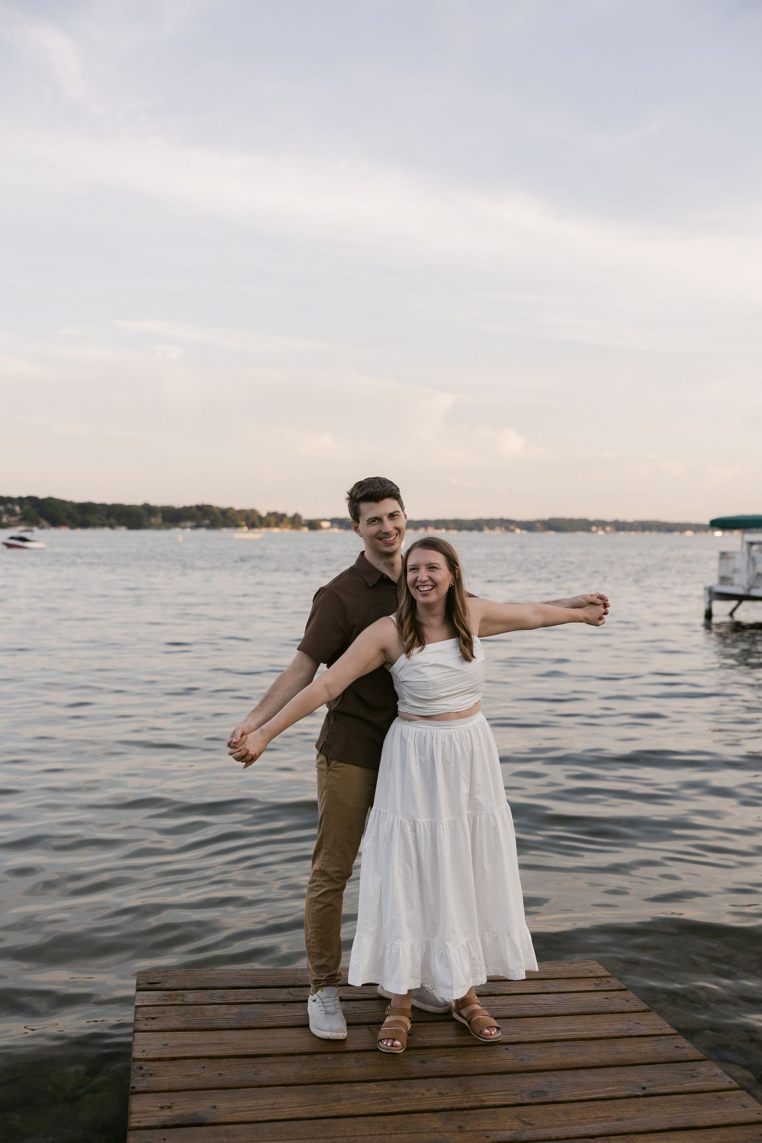 Two people dancing on a lakeside dock at dusk with boats visible in the distance.