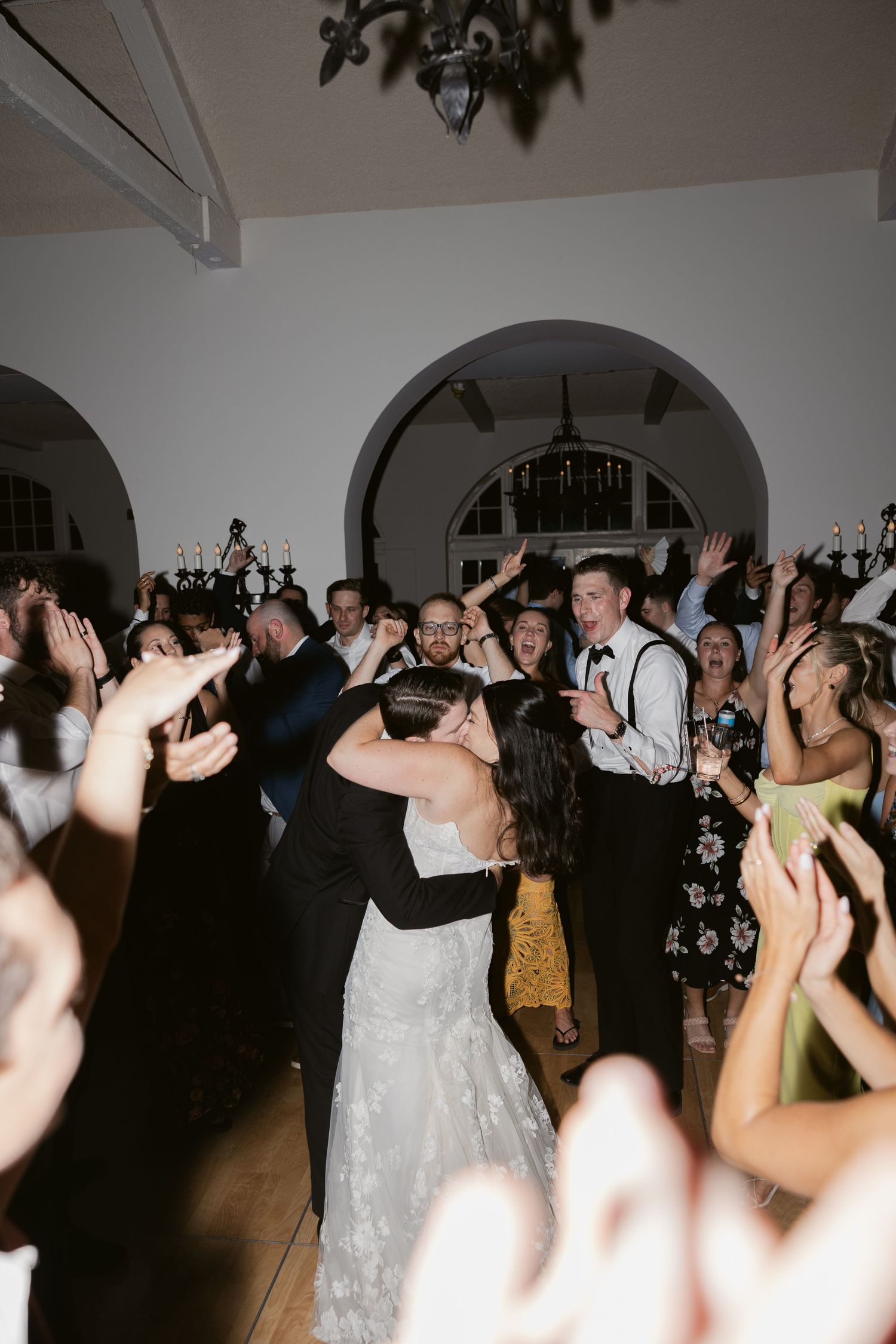 Wedding reception dance floor filled with guests dancing and celebrating in a white-walled venue with arches.