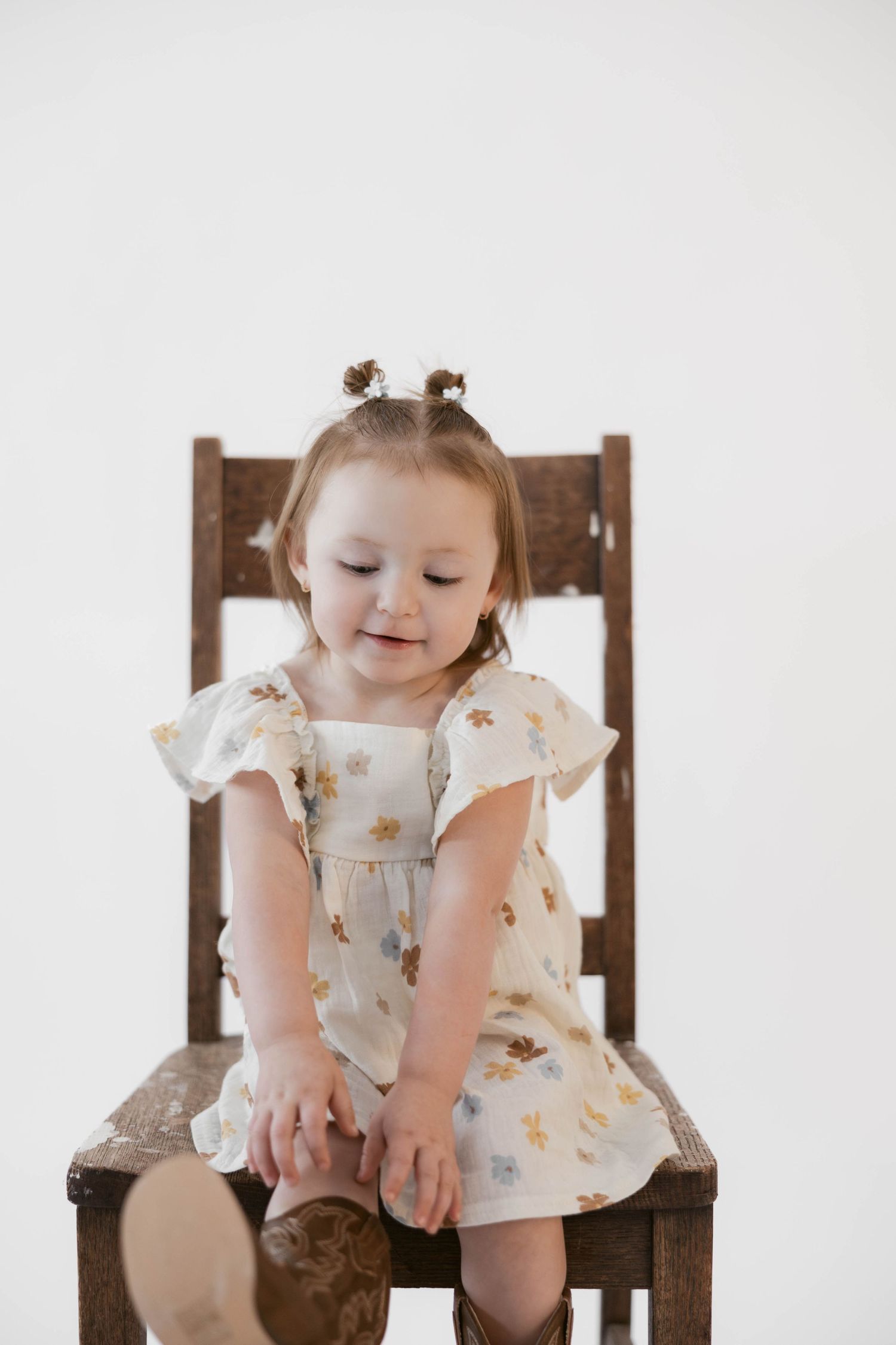 Cheerful toddler wearing floral dress looks down while seated on wooden chair.