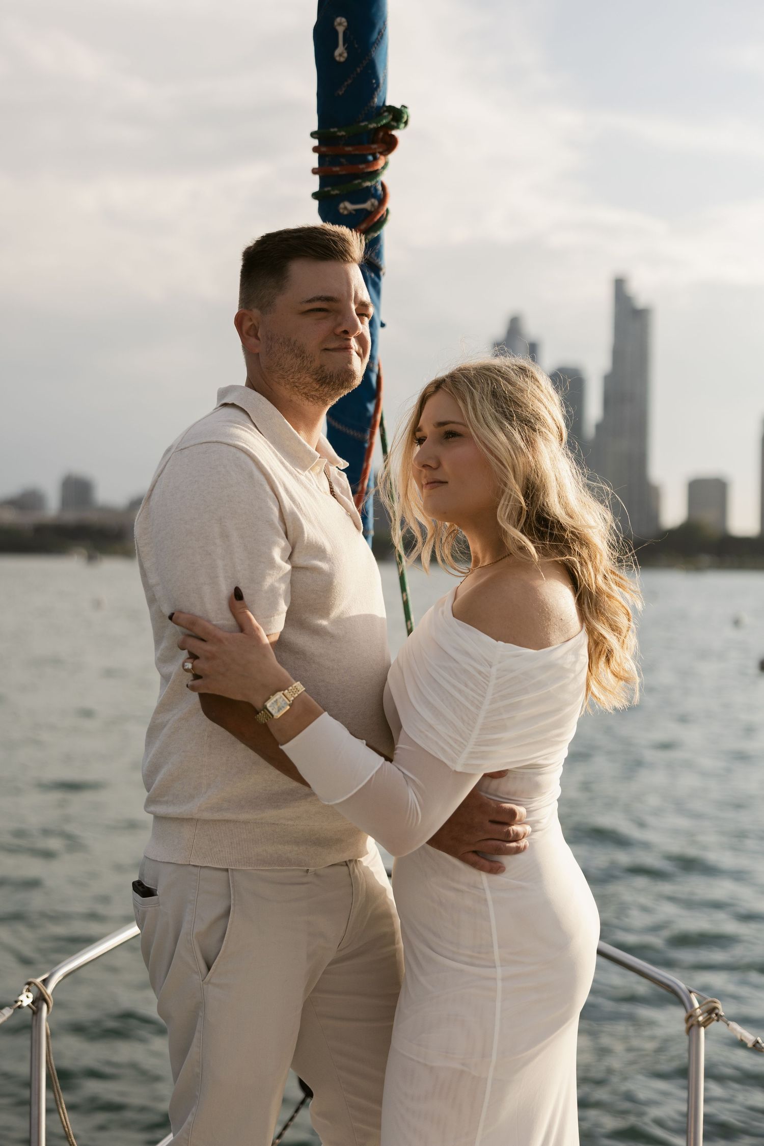 A couple in white outfits shares an intimate moment on a boat dock with a cityscape and water in the background.