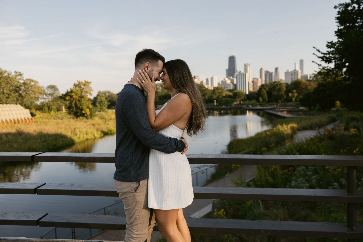 A couple embraces on a bridge with the Chicago skyline visible across the water during golden hour.