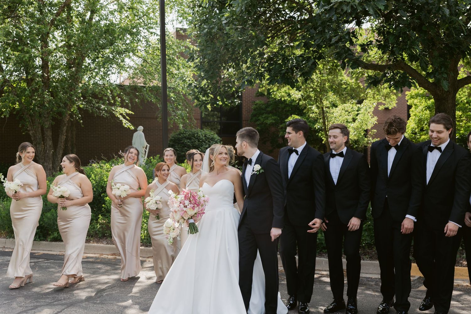 A wedding party in pale pink bridesmaid dresses and classic black tuxedos poses outdoors on a sunny day.