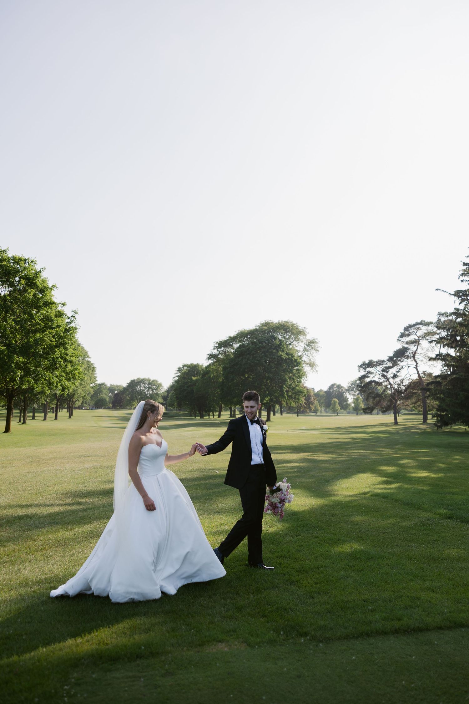 Newlyweds walking hand in hand across a sweeping golf course landscape.