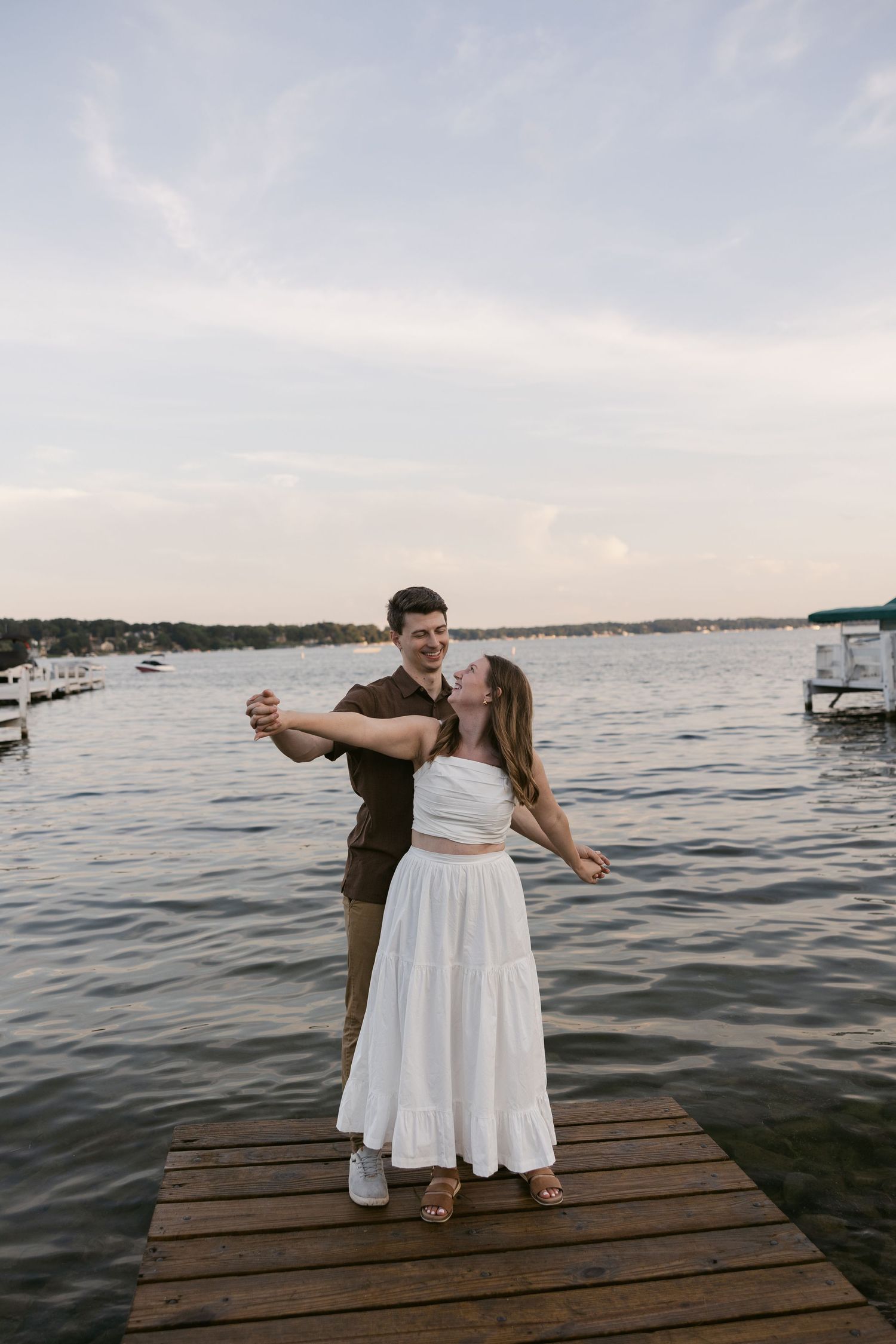 A couple dances playfully on a wooden dock during sunset with a lake and boats in the background.