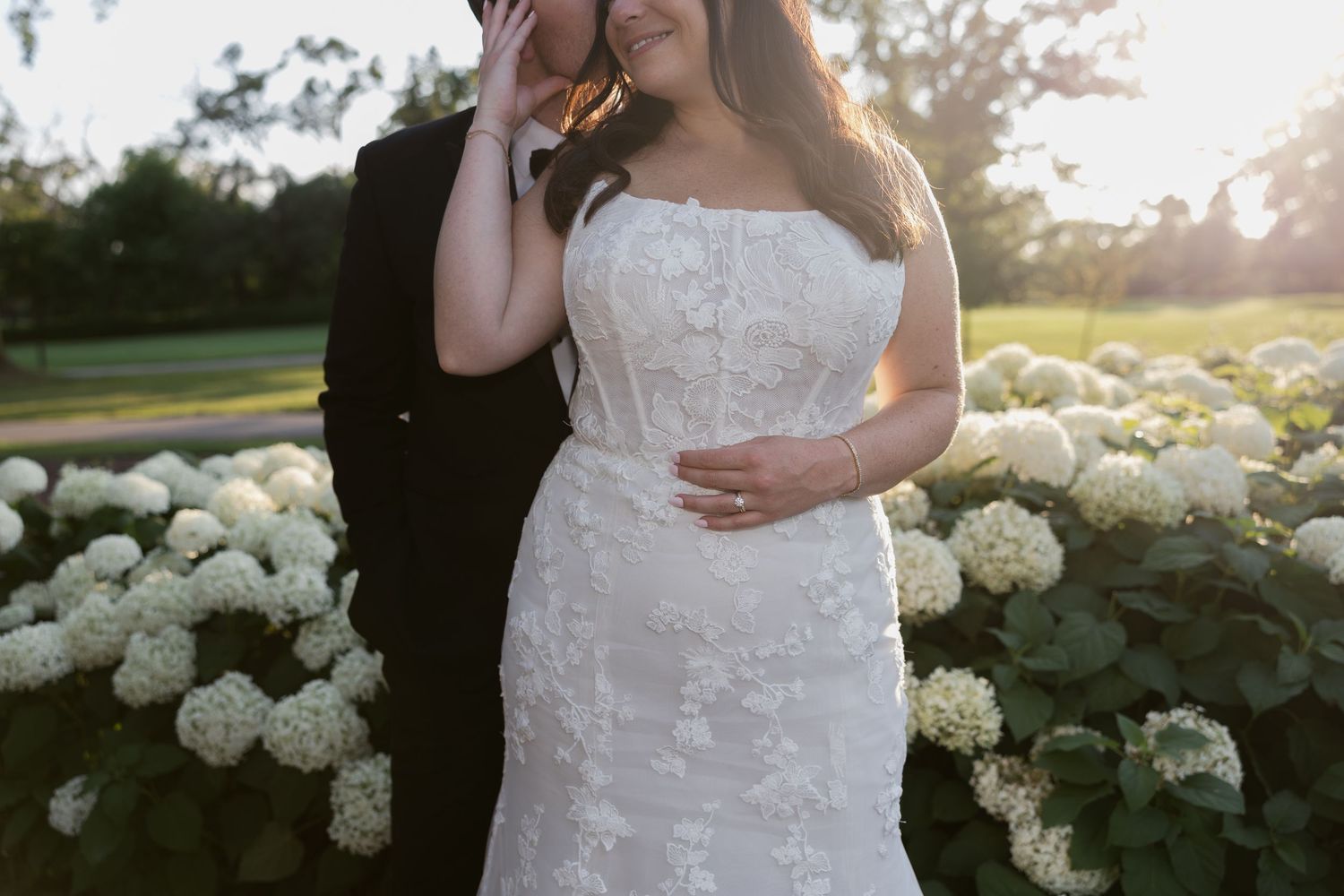 Bride in lace wedding dress poses among blooming white hydrangea bushes at sunset.