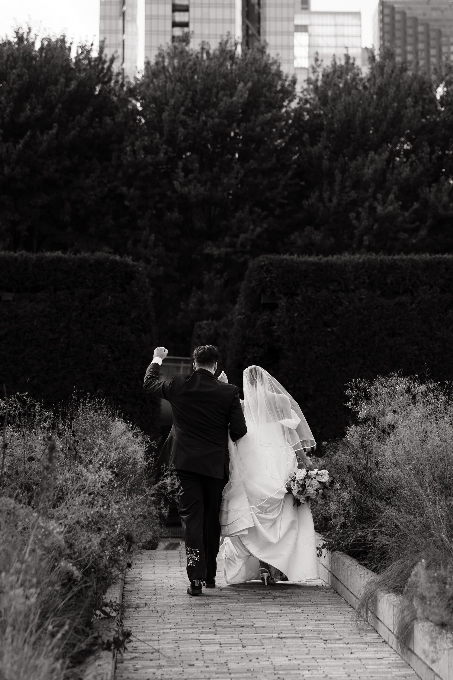 A wedding couple walks along a garden path in black and white, with architectural elements in the background.