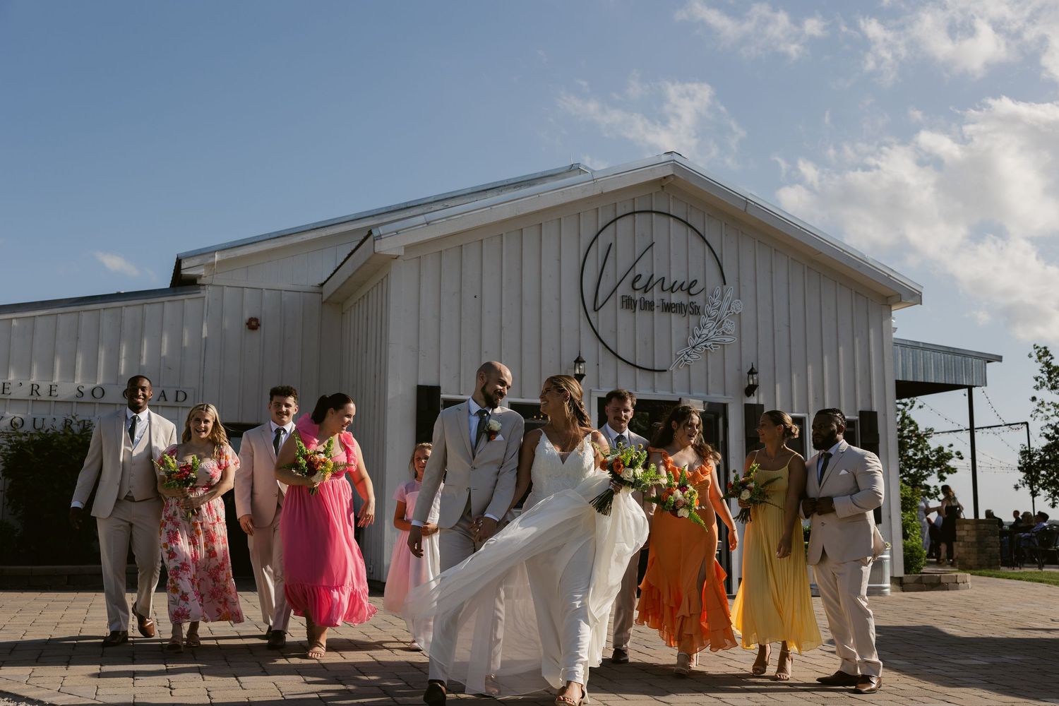 A wedding party in colorful attire walks together outside a white barn venue on a sunny day.
