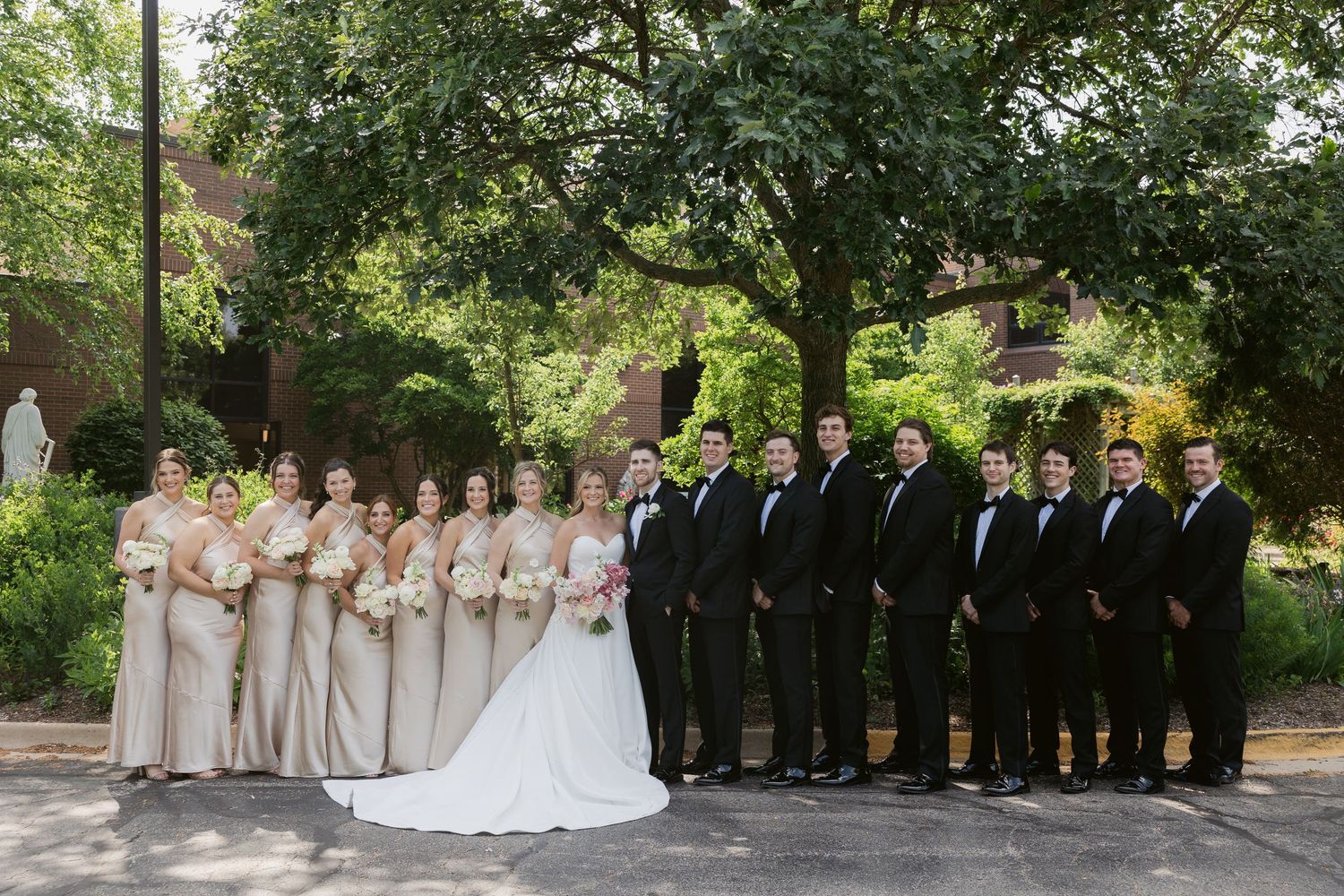 A full wedding party in black and champagne attire poses formally outdoors under trees.