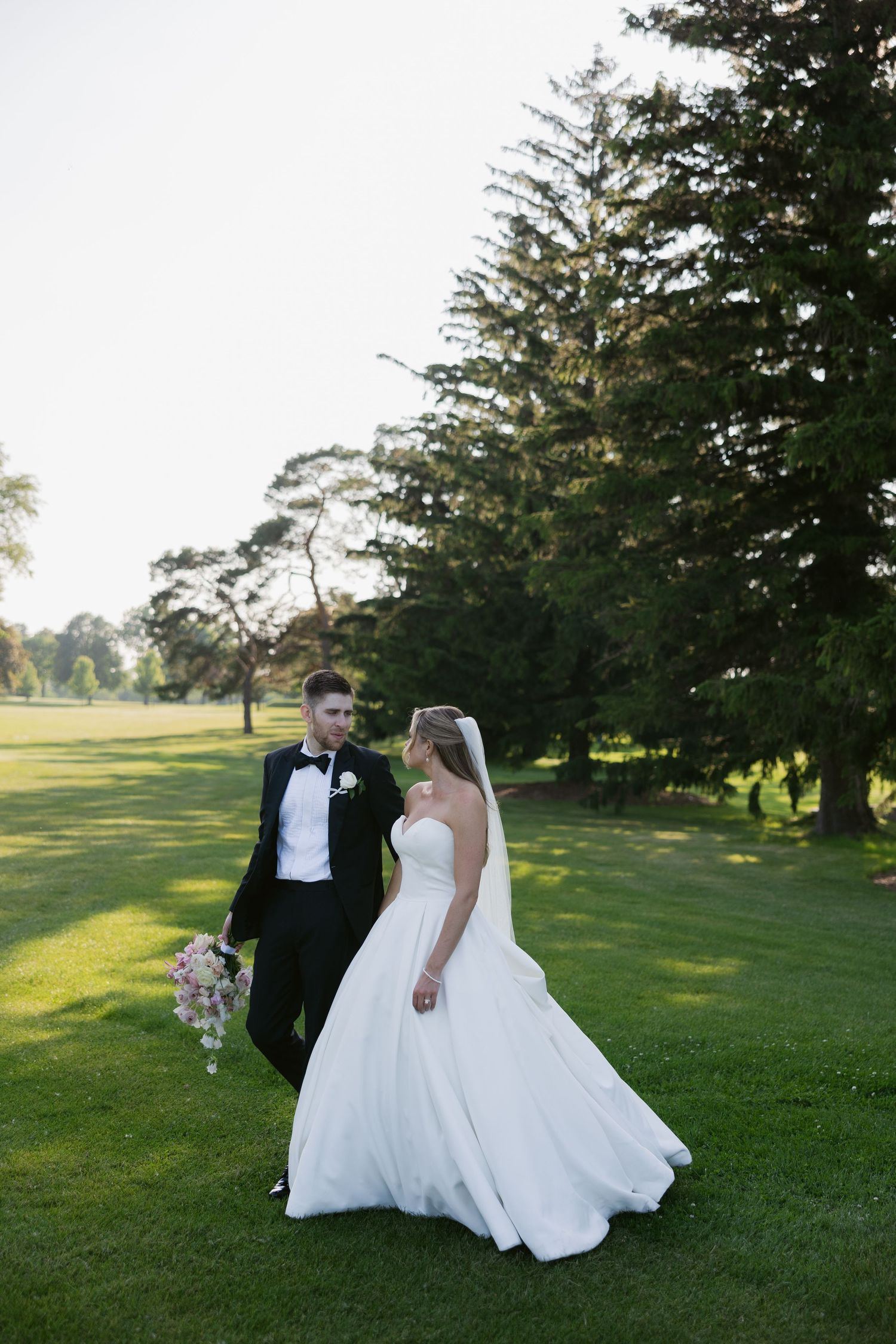 Wedding couple walks on lush green lawn with towering evergreen trees in the background.