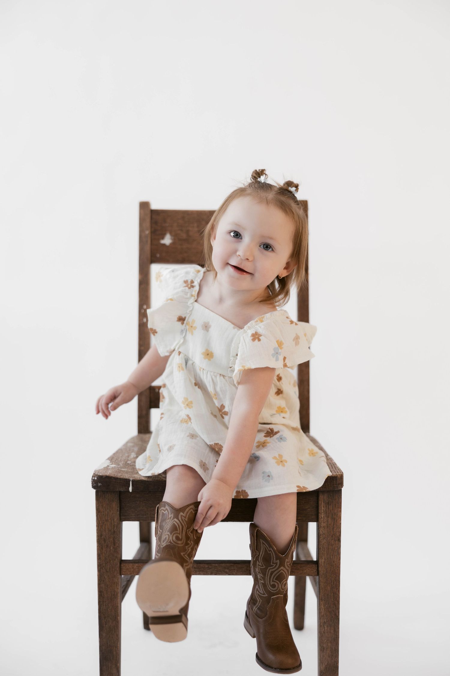 Happy toddler in floral dress and brown boots posing playfully on wooden chair.