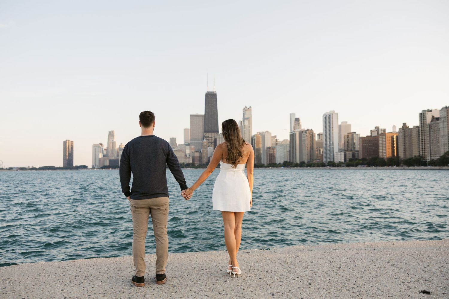 A couple walks along the shoreline with the Chicago skyline silhouetted against a bright evening sky.