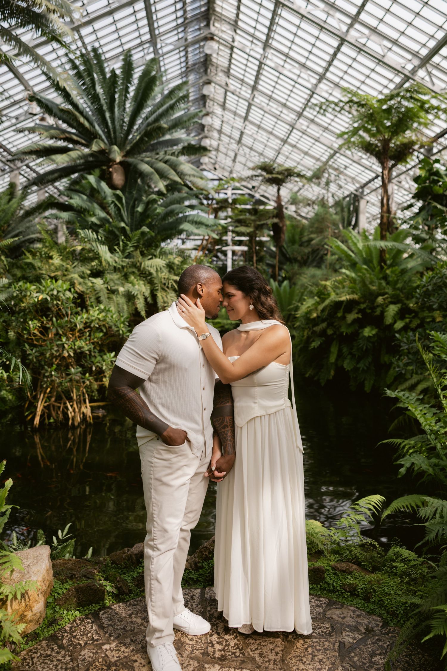 A tender moment shared between a couple in white attire surrounded by tropical plants in a greenhouse setting.
