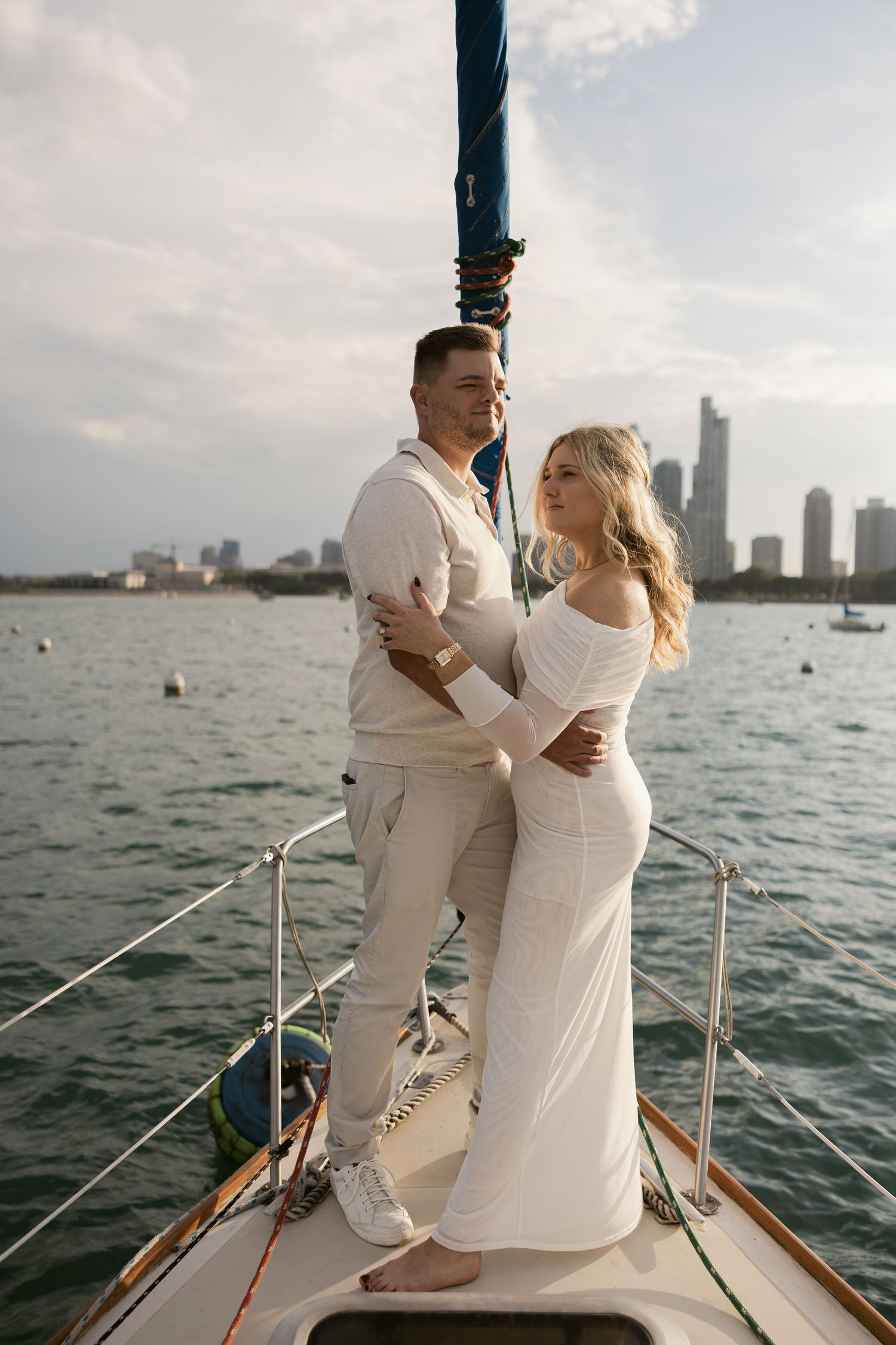 A romantic couple embraces on the deck of a sailboat with a waterfront cityscape visible in the distance.