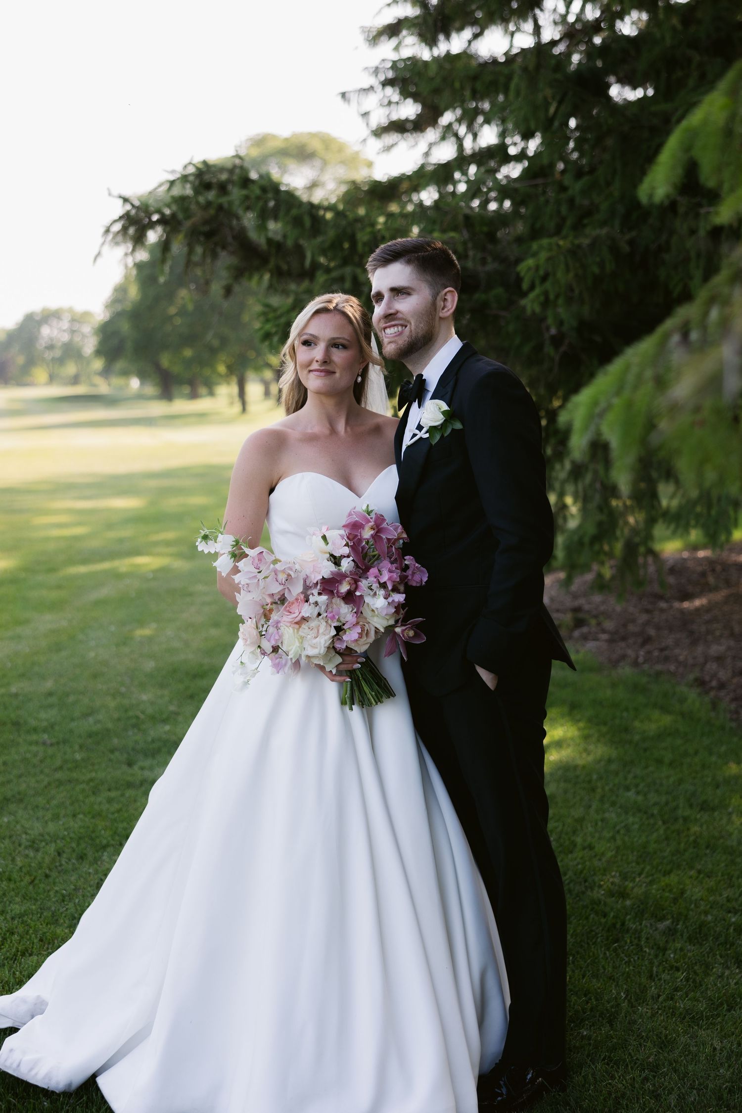 Elegant wedding couple poses outdoors with pink bouquet under evergreen trees at sunset.