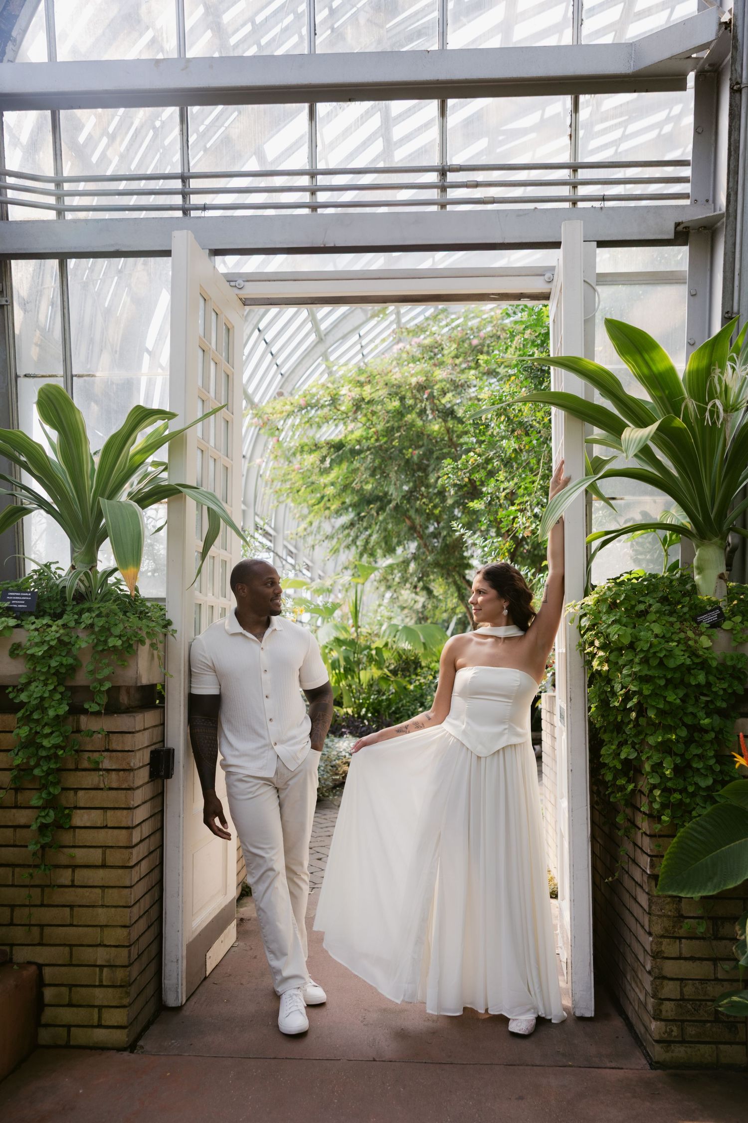 Newlyweds share a joyful moment while walking through a sunlit greenhouse filled with greenery and plants.