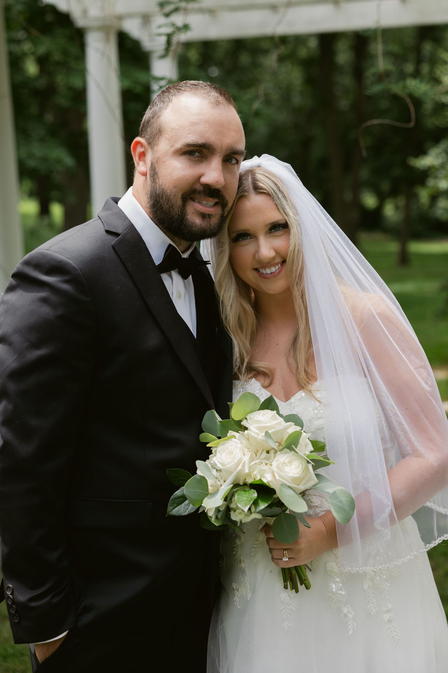 A joyful couple on their wedding day standing together outdoors, with the bride holding a bouquet of white roses.