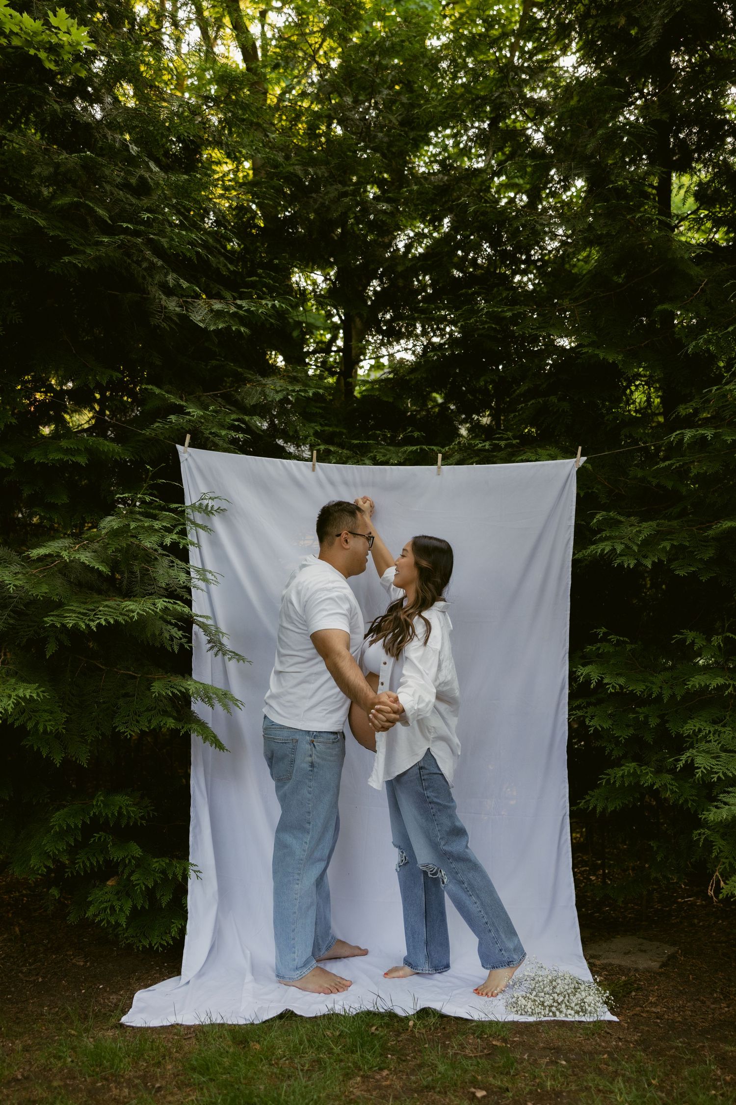 A tender maternity photoshoot against a white backdrop in a lush green forest setting.