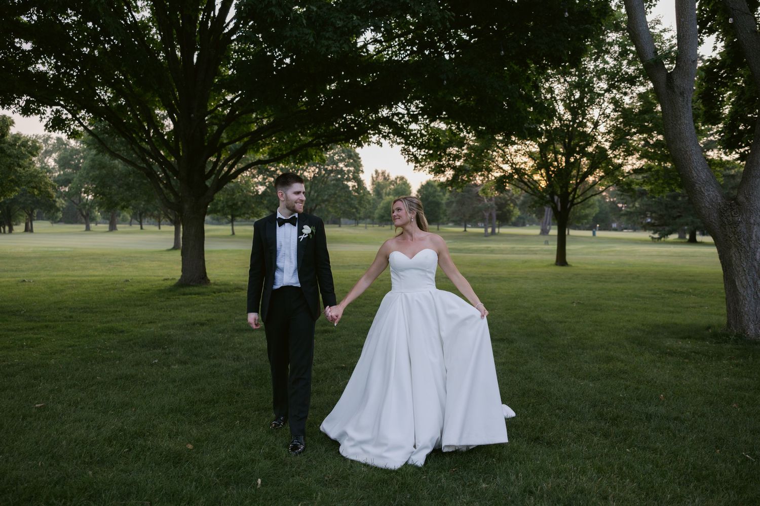 Newlywed couple walking together on grassy lawn during golden hour.