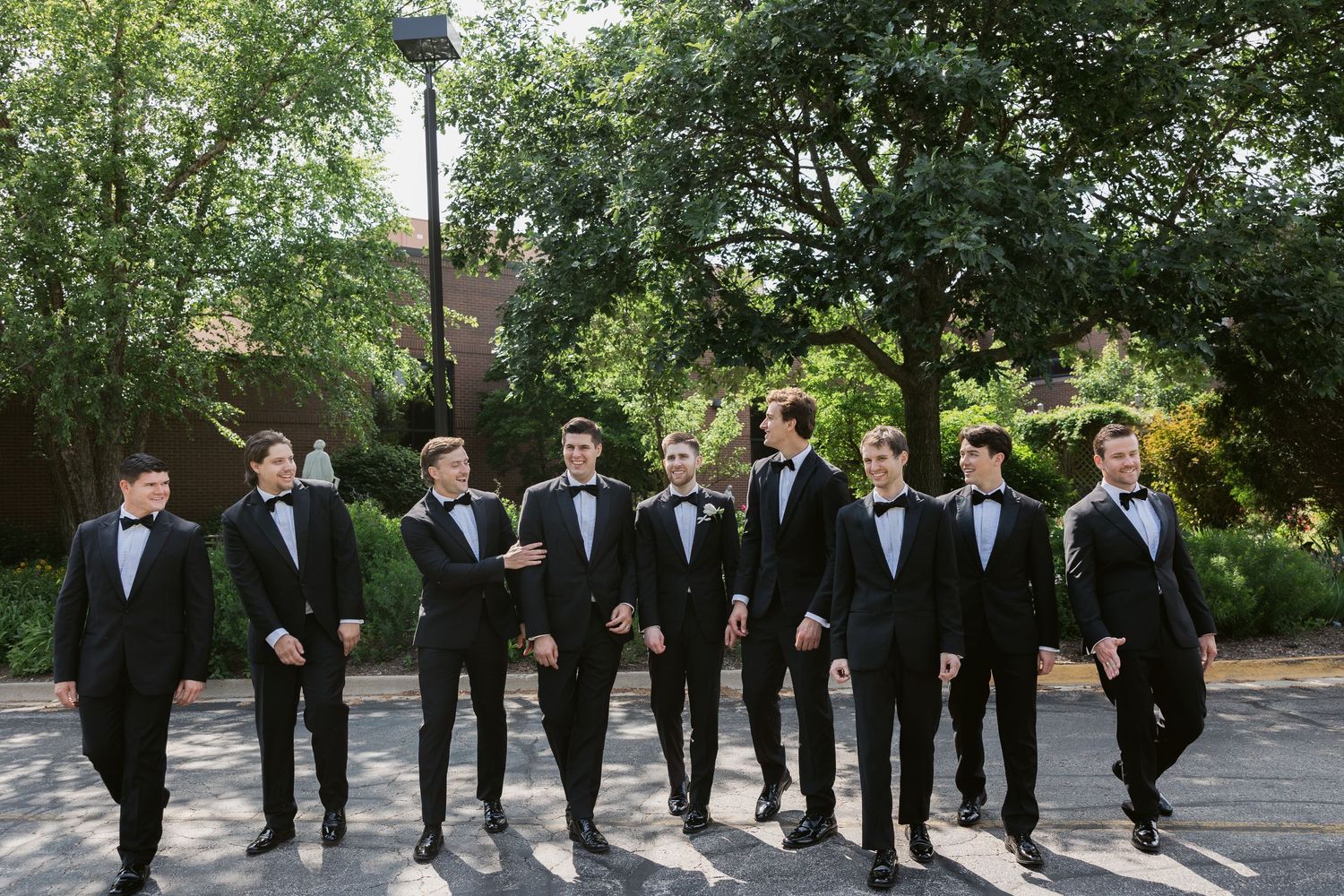 Wedding party members in black tuxedos posing in a line on a garden pathway.