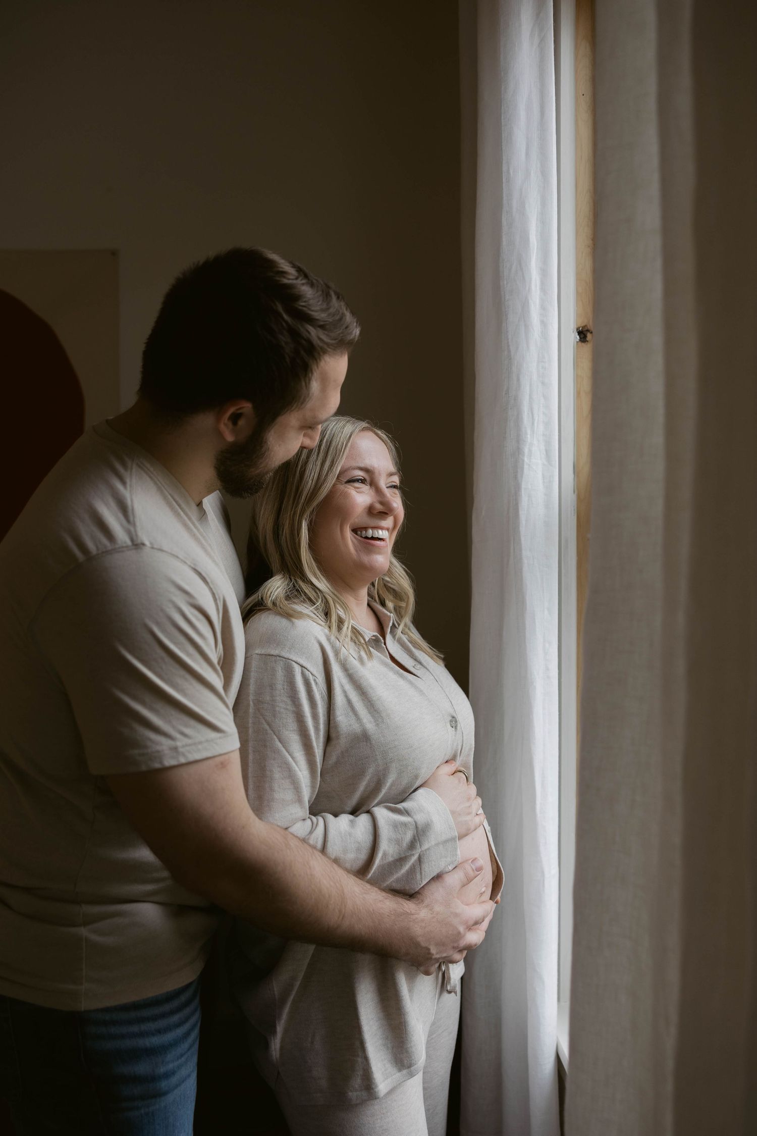 A romantic maternity photo of an expectant couple sharing a tender moment by a window in natural light.