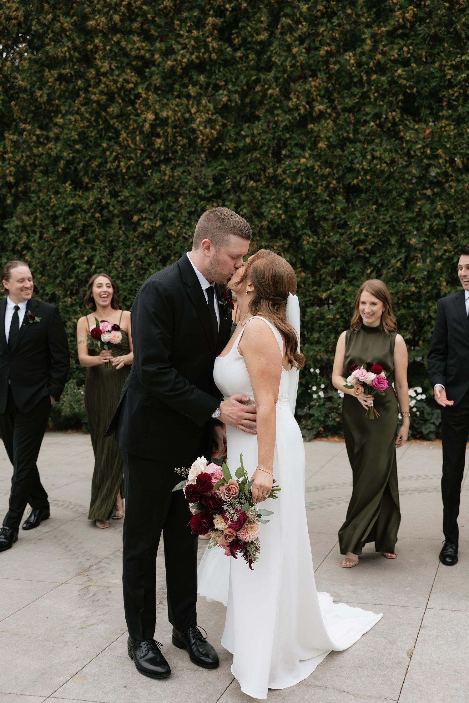 A wedding party in formal attire stands together on a stone patio with green hedges in the background.